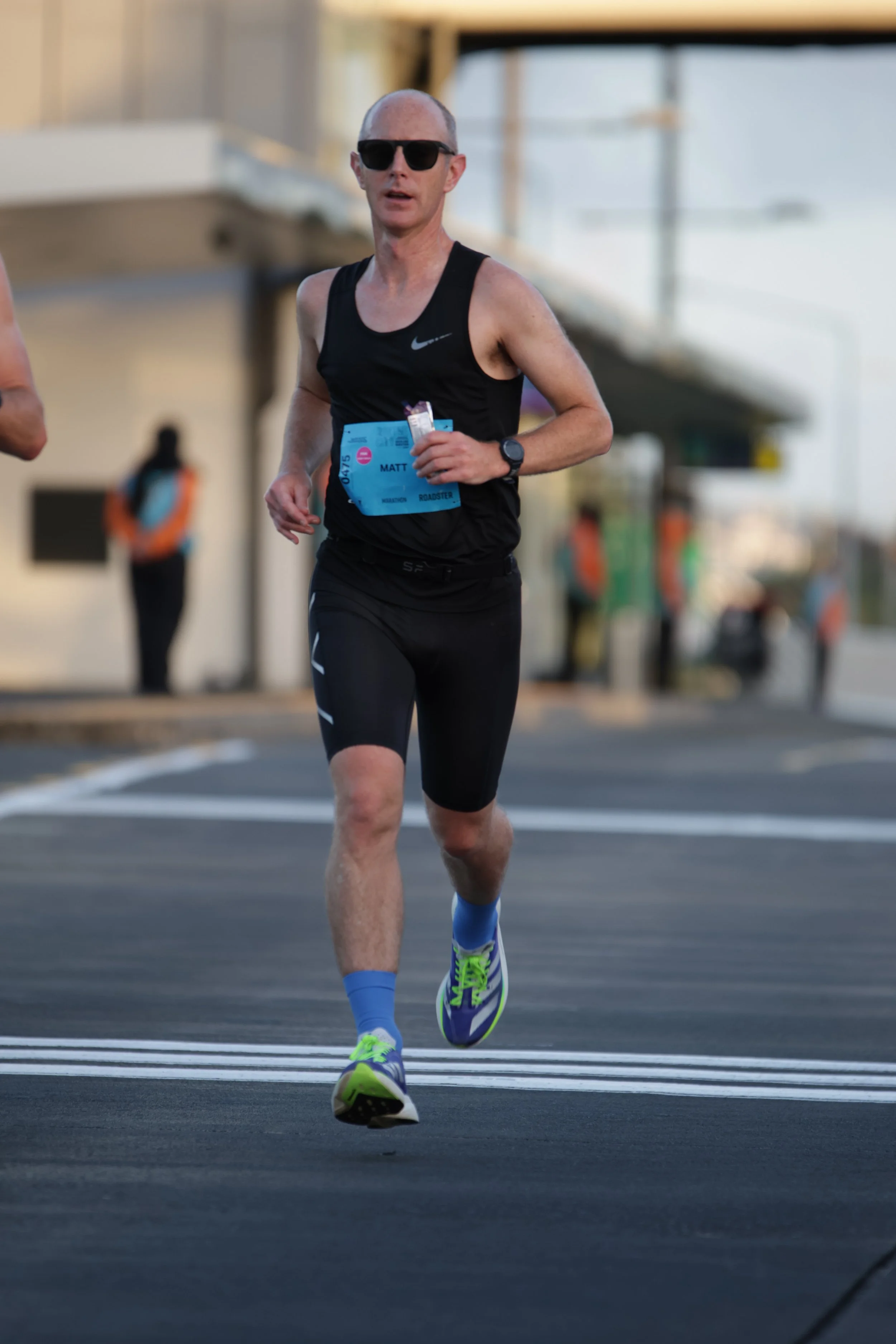 A man running in a marathon or race wearing sunglasses, a black Nike tank top, black shorts, blue compression socks, and bright yellow and purple running shoes. He has a race bib with the name 'Matt' on it.