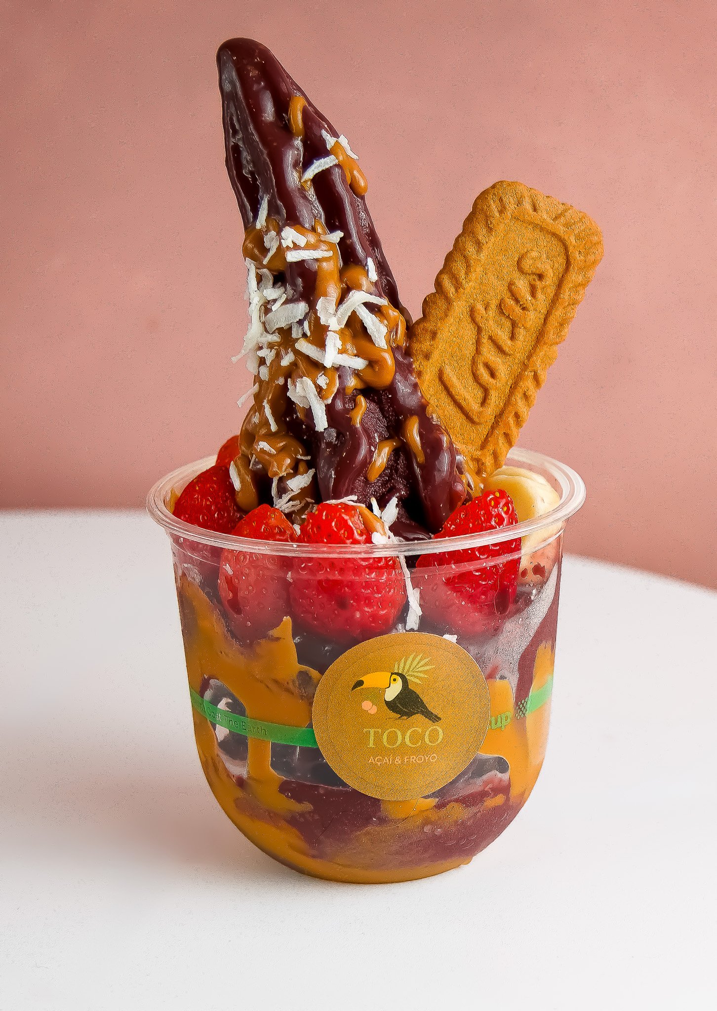 A colorful cup filled with strawberries, bananas, and other fruit, topped with a thick chocolate and caramel drizzle, shredded coconut, and a cookies and cream biscuit.