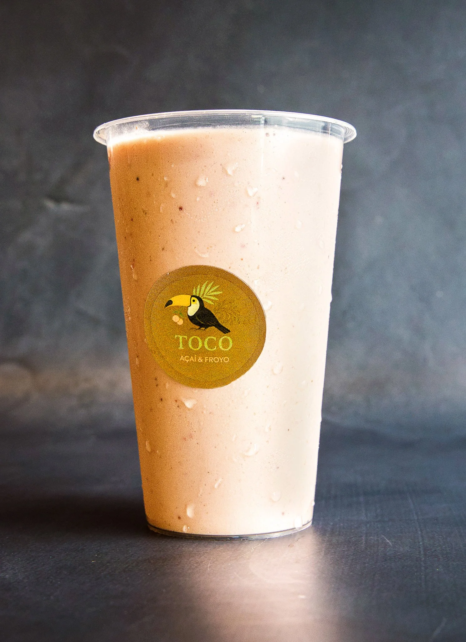 A plastic cup filled with a creamy, pink-colored acai frozen beverage with ice particles visible on the outside. The cup has a yellow sticker with a toucan logo and the words "TOCO ACÁI & FROYO." The background is black and the cup is sitting on a da