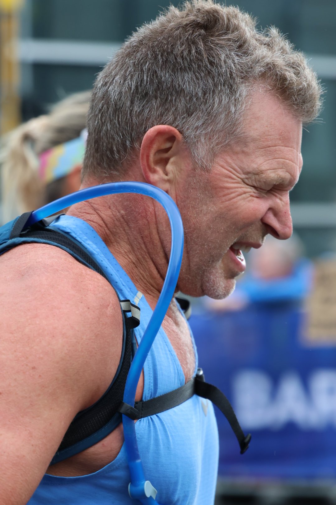 Older man with short gray hair, sweating and smiling, wearing a blue tank top and hydration gear, likely participating in a race or outdoor event.