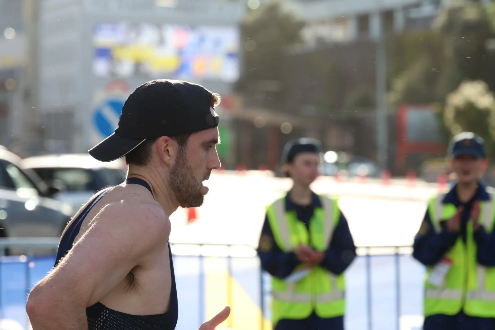 A male runner wearing a black cap and sleeveless shirt runs past two women in reflective vests who are standing near traffic cones during an outdoor race or marathon.