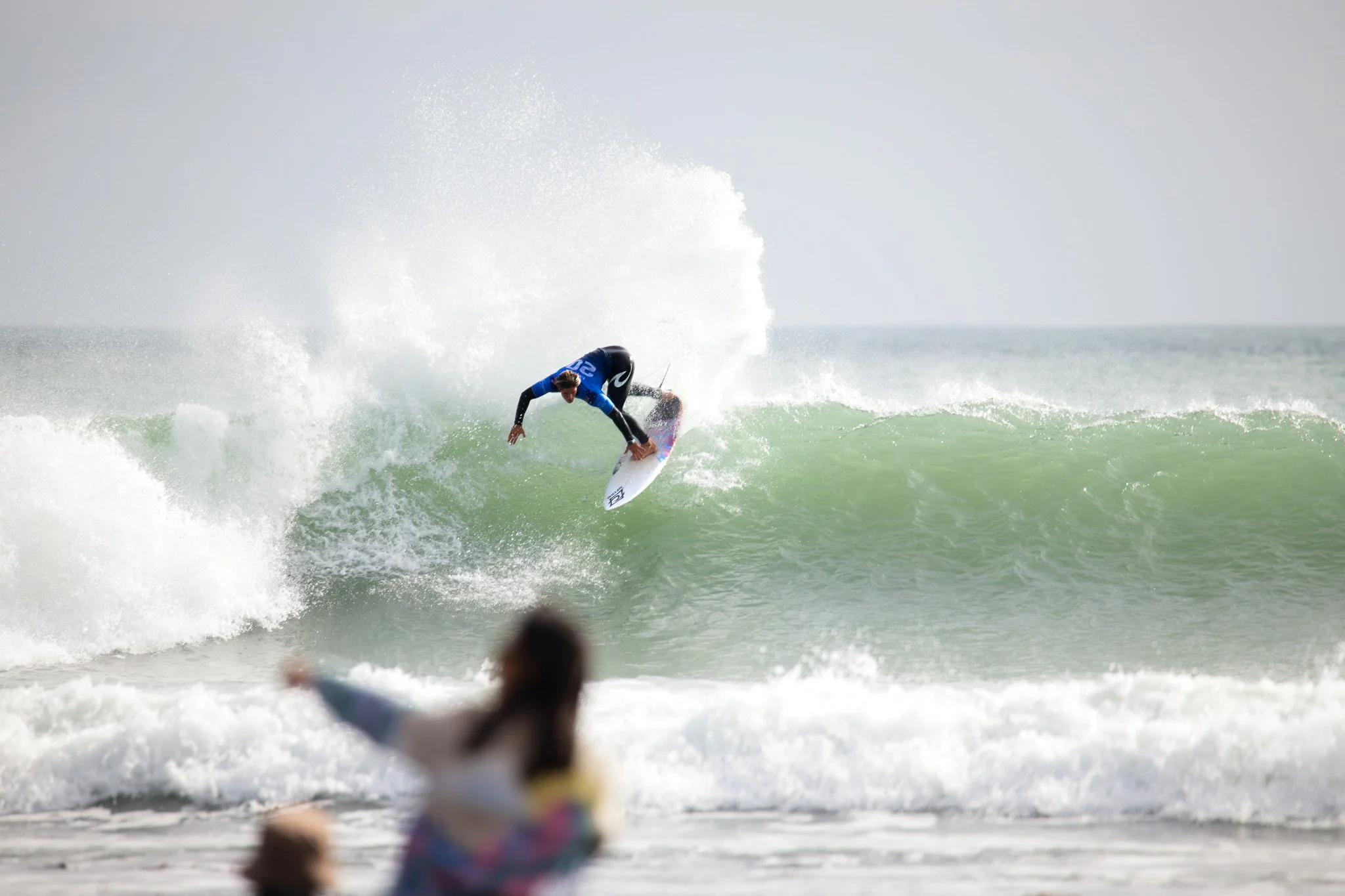 Surfer riding a wave in the ocean during daytime, with spectators in the foreground.