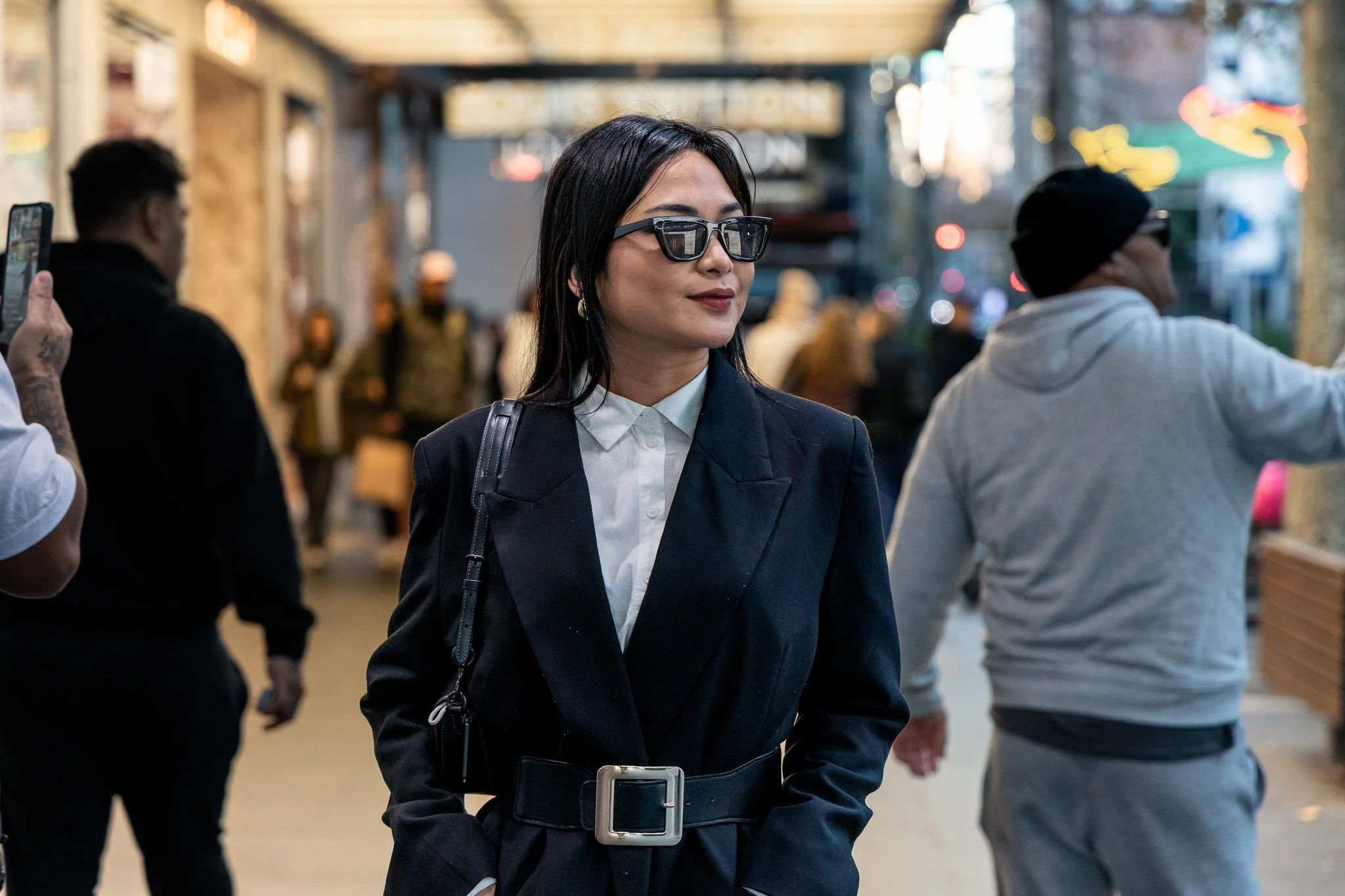 A woman in a dark coat and sunglasses walking outdoors in an urban setting, with other people around her and blurred city lights in the background.