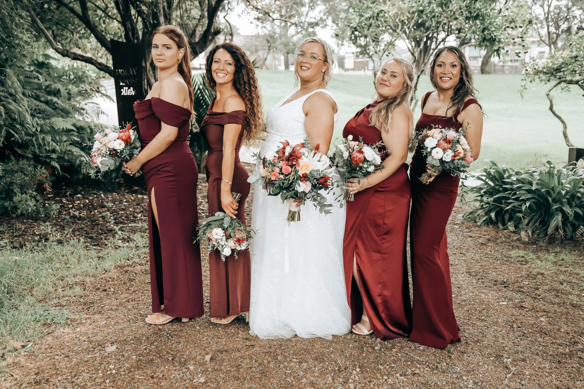 A group of six women dressed in formal attire, standing outdoors among trees, holding bouquets, with a mix of smiles and neutral expressions.