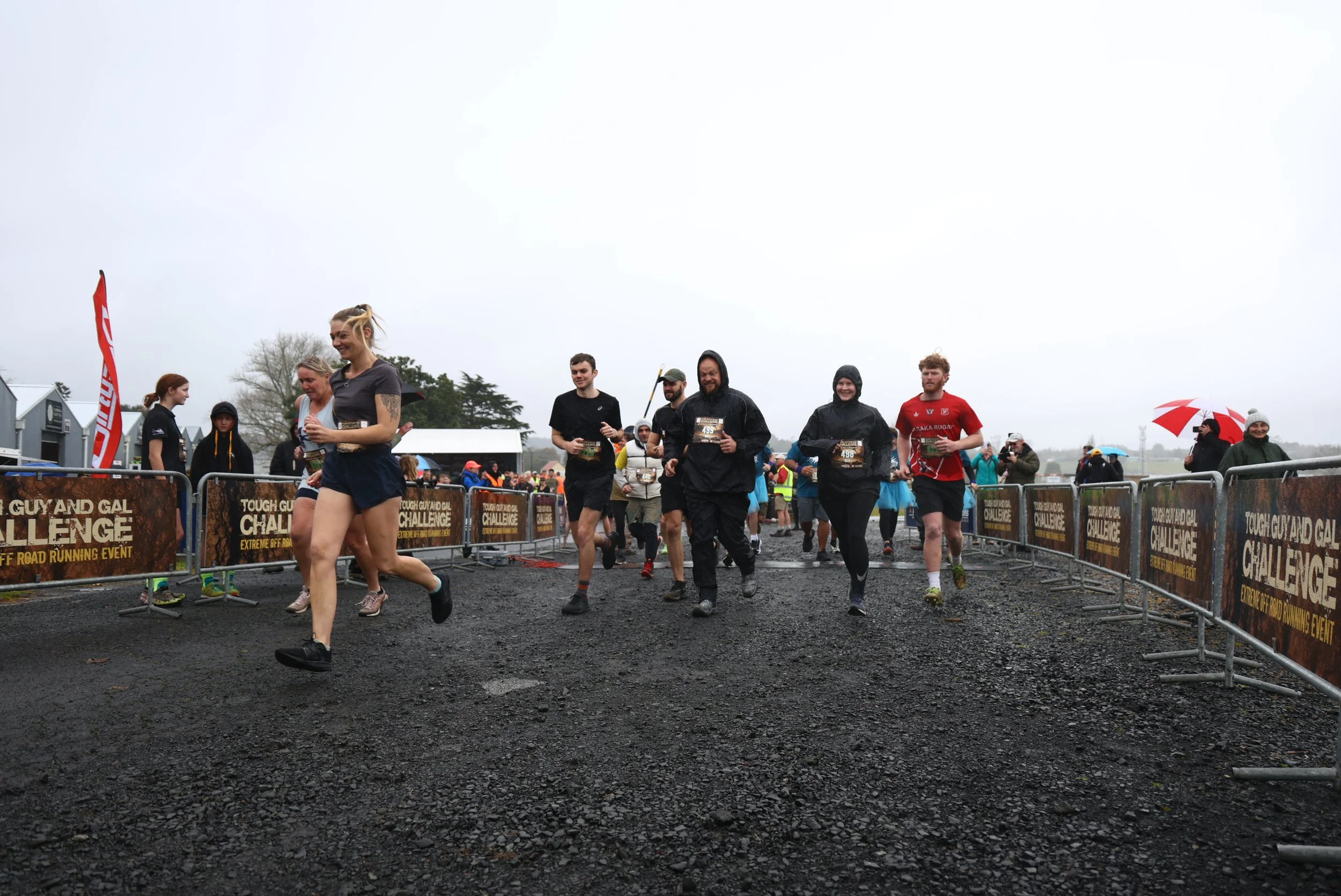 People participating in an outdoor race event on a rainy day, with some wearing rain jackets and holding umbrellas, crossing the finish line.