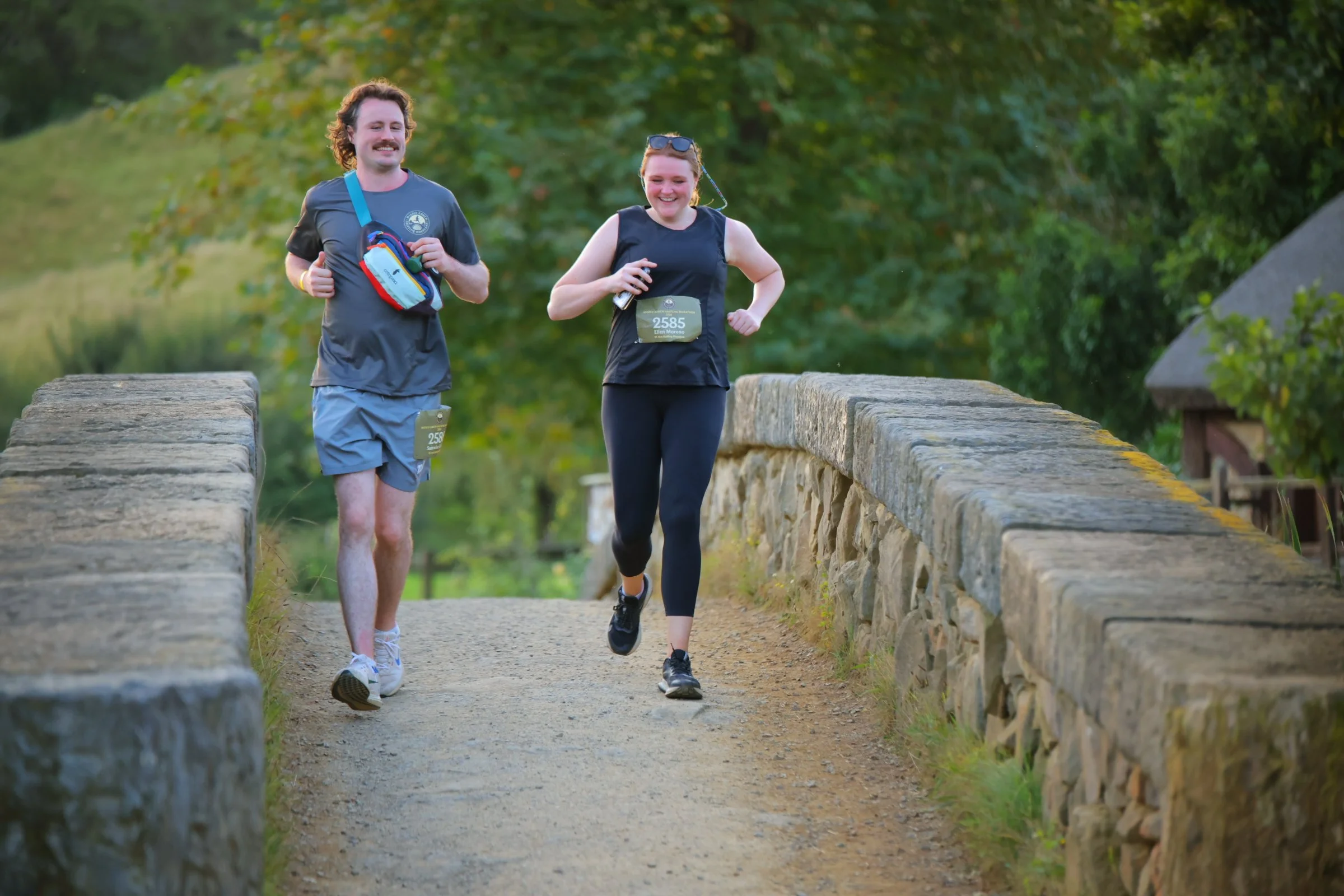 A man and a woman running on a stone bridge in a park, both wearing athletic clothing and race bibs, smiling.