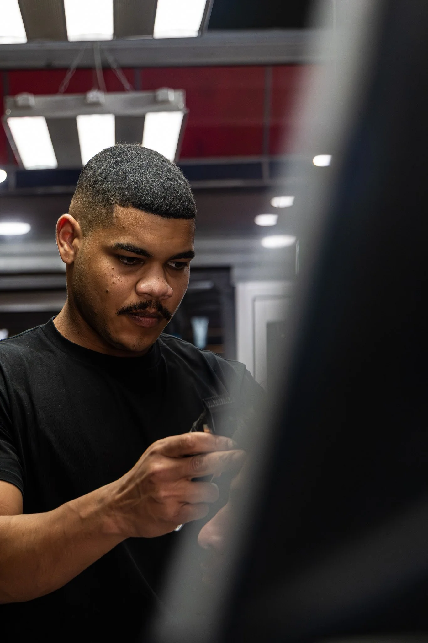 A man with short dark hair, a mustache, and light skin, wearing a black shirt, is looking at his phone while standing in an indoor setting. There are bright ceiling lights and a red wall in the background.