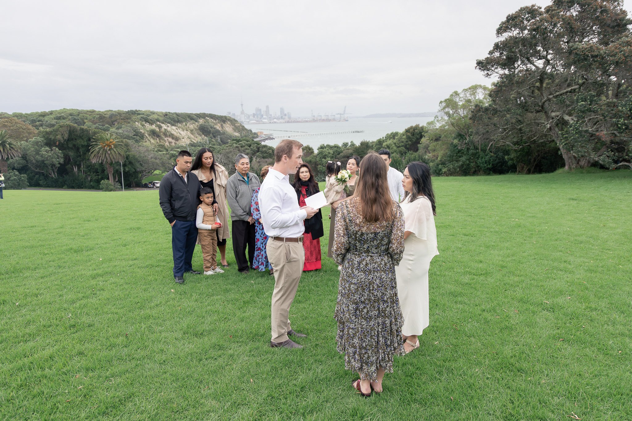 A group of people participating in a wedding ceremony outdoors on a grassy area with a city skyline and water in the background. The officiant is reading from a book, with the bride and groom standing in front of him, while friends and family watch.
