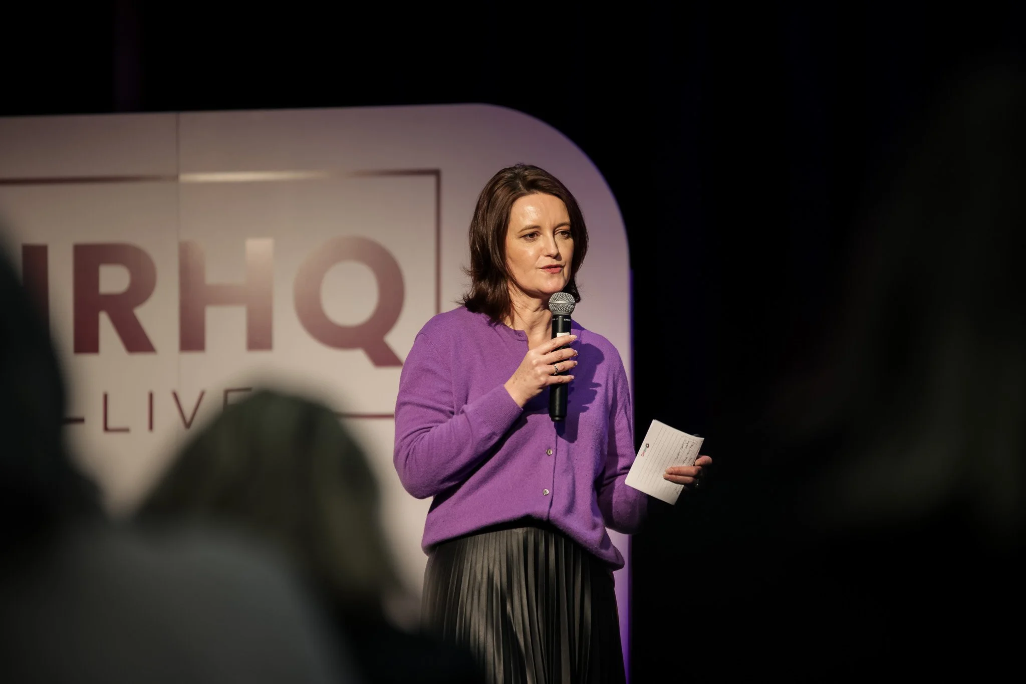 A woman with shoulder-length brown hair, wearing a purple cardigan, holding a microphone and some papers, speaking on stage at an event.