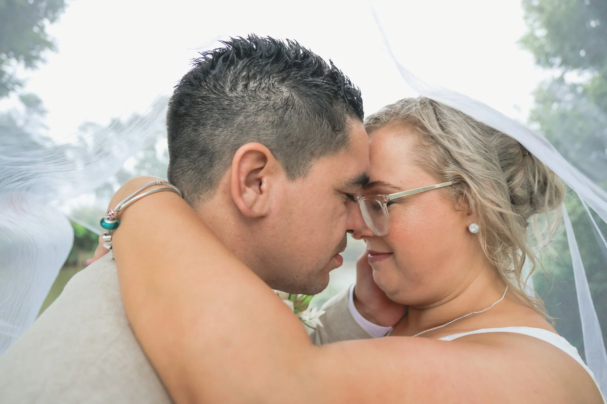A close-up of a couple with their foreheads touching, eyes closed, sharing an intimate moment outdoors under a white fabric covering, with trees and cloudy sky in the background.