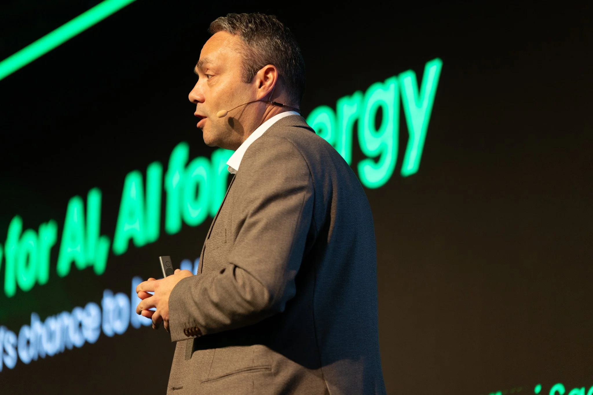 A man in a gray suit holding a remote control on stage with green neon text in the background.