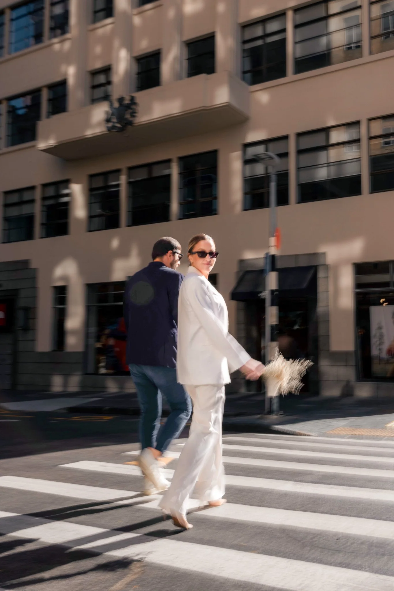Two people crossing the street at a crosswalk in an urban area. The woman is dressed in a white suit and holding a bouquet, while the man is in a dark jacket and jeans. The buildings in the background are modern with multiple windows.