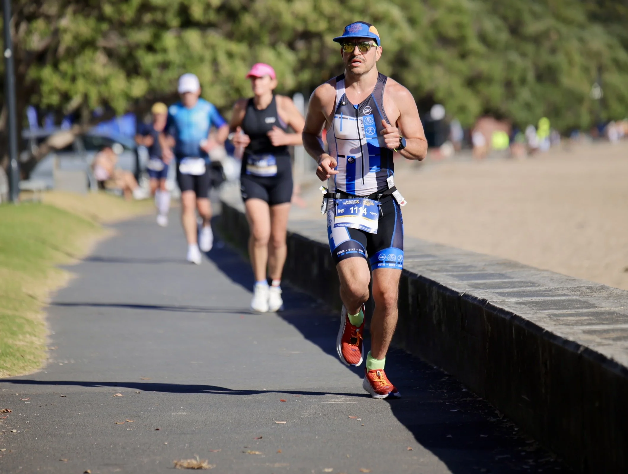 A man running outdoors in a race, wearing a blue and black athletic outfit, bright orange running shoes, sunglasses, a blue cap, and a race bib with the number 1114. Several other runners are visible in the background on a paved path lined with green