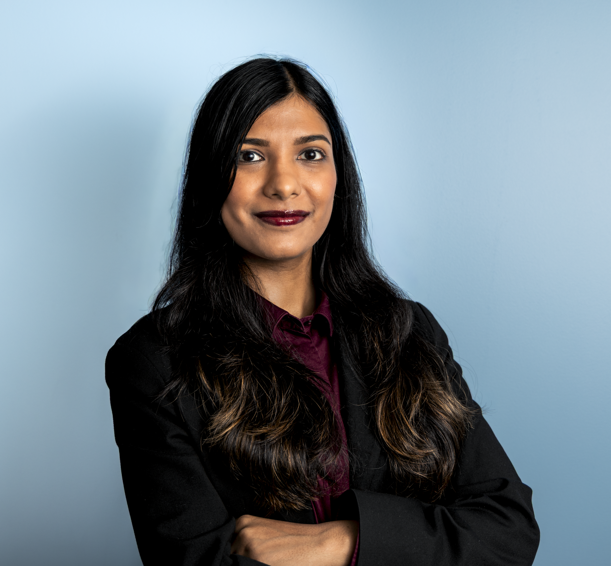 A woman with long, dark hair, wearing a dark blazer and a maroon shirt, standing against a light blue background, with arms crossed and smiling slightly.