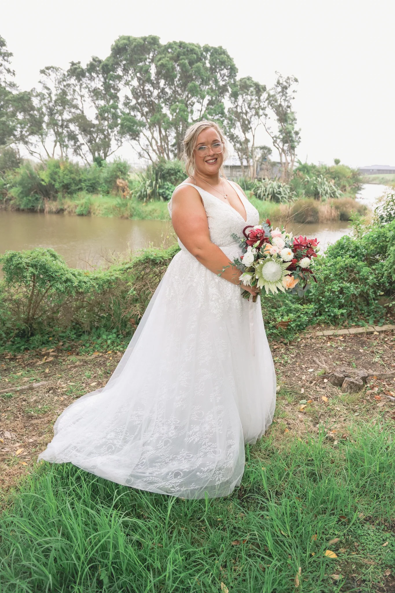 A woman in a white wedding dress holding a bouquet of flowers, standing outdoors by a river with lush green trees and bushes in the background.