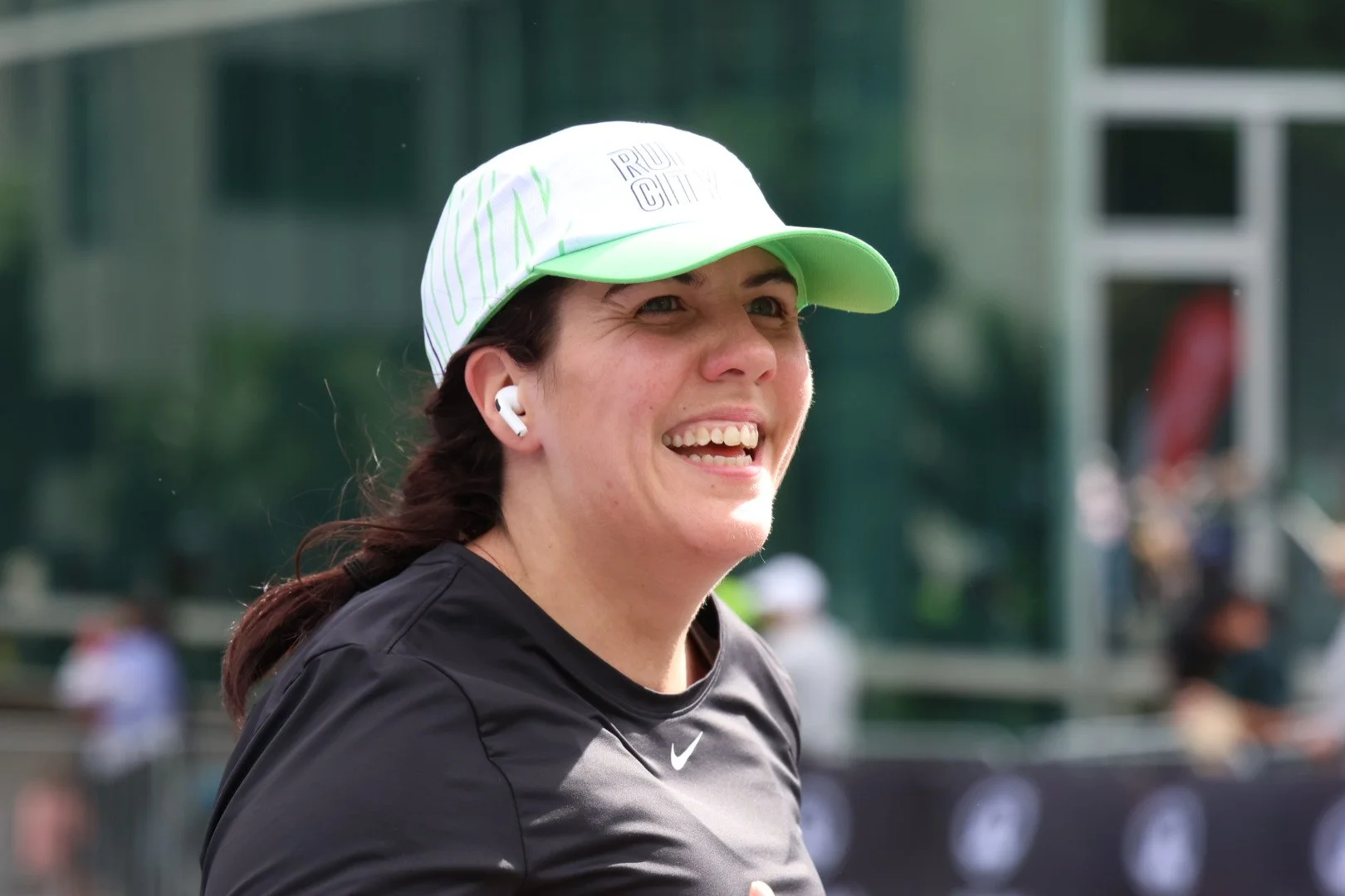 A woman smiling, wearing a white sports cap with green accents, AirPods, and a black Nike shirt, outdoors at a sporting event.
