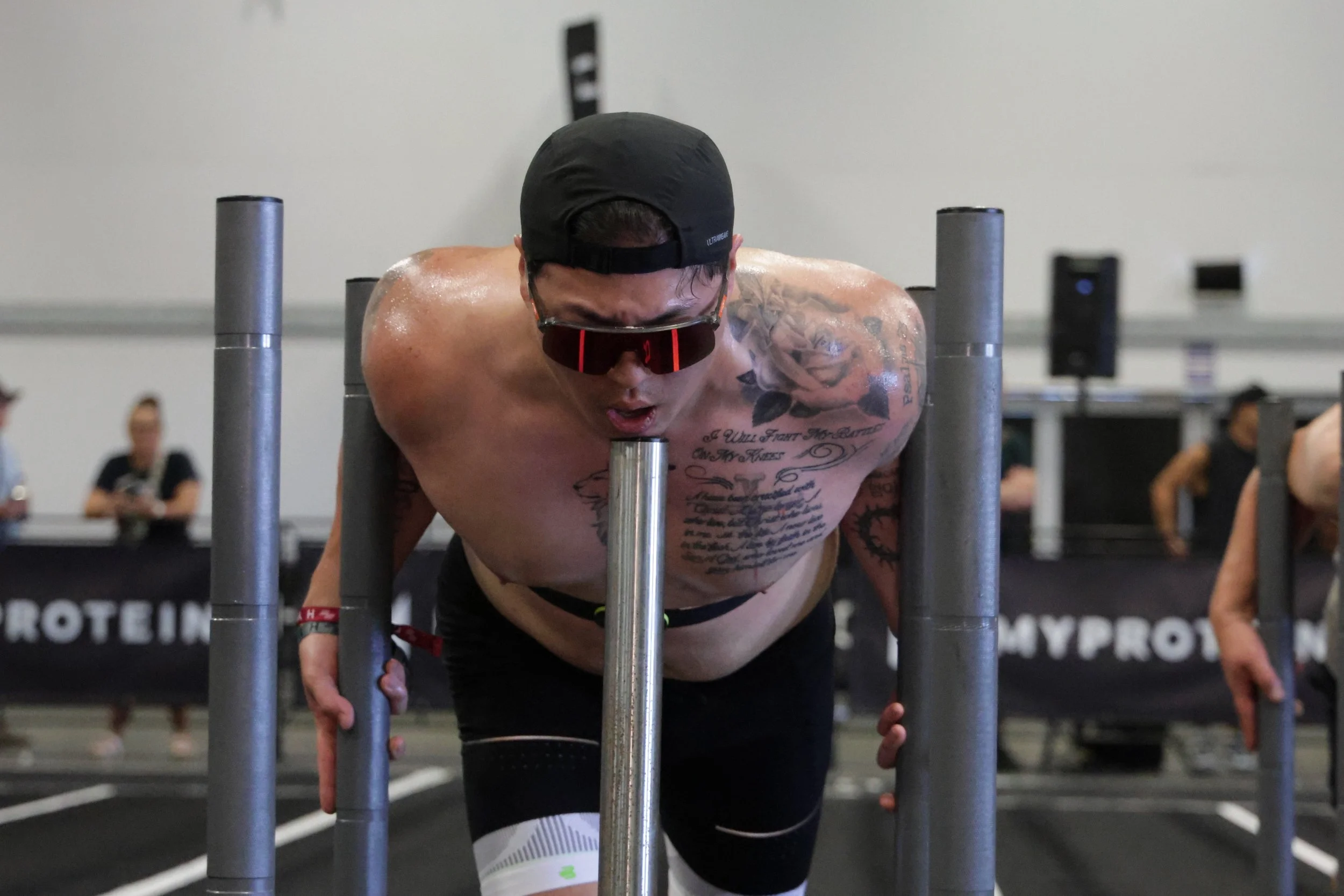 A shirtless man with tattoos, wearing a black cap and sunglasses, is exercising with a barbell at a gym.