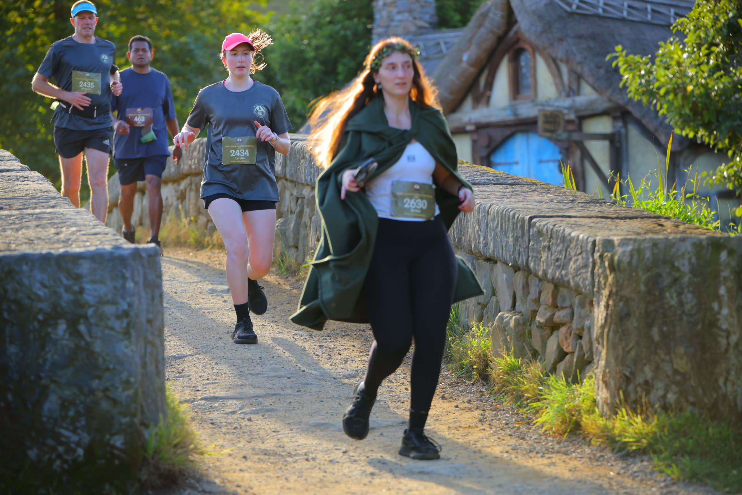 Group of people running outdoors along a stone pathway with greenery and a stone building in the background.