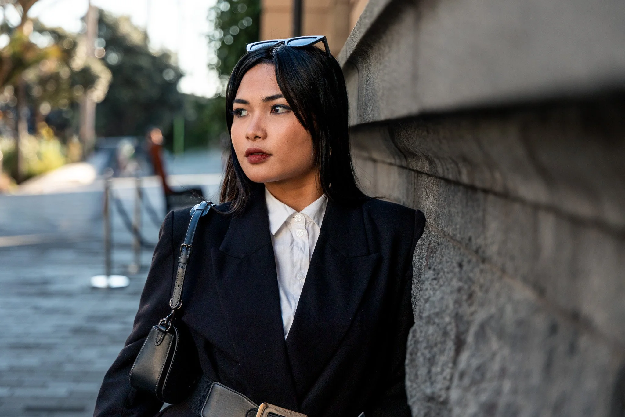 A woman with black hair and sunglasses on her head leans against a stone wall on a city street, wearing a black blazer and white shirt, with a black shoulder bag.