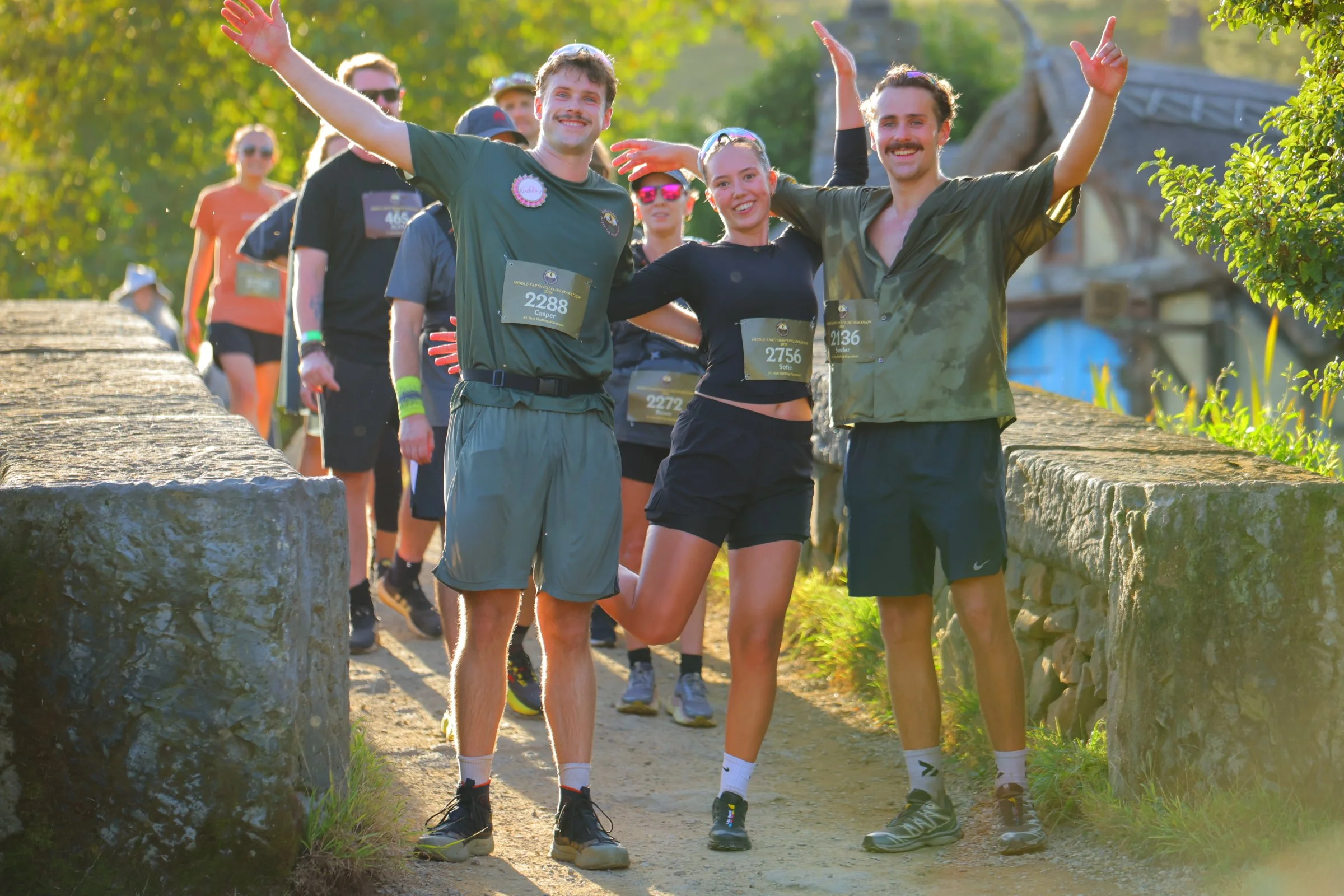 People crossing a stone bridge during a running event, smiling and waving, wearing athletic gear and bibs with numbers, surrounded by greenery.