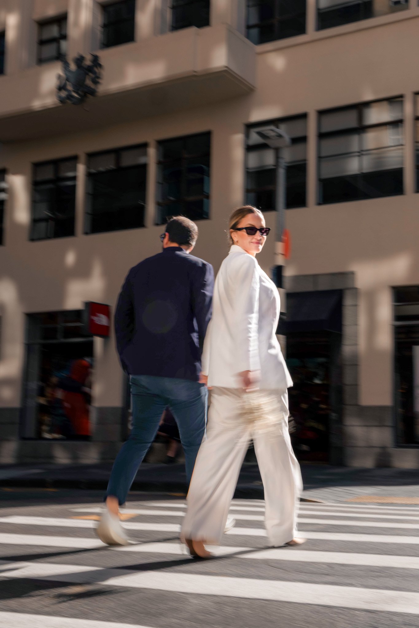 Woman in white suit and sunglasses walking on crosswalk with man in dark jacket and glasses behind her, urban background with buildings and shadows.