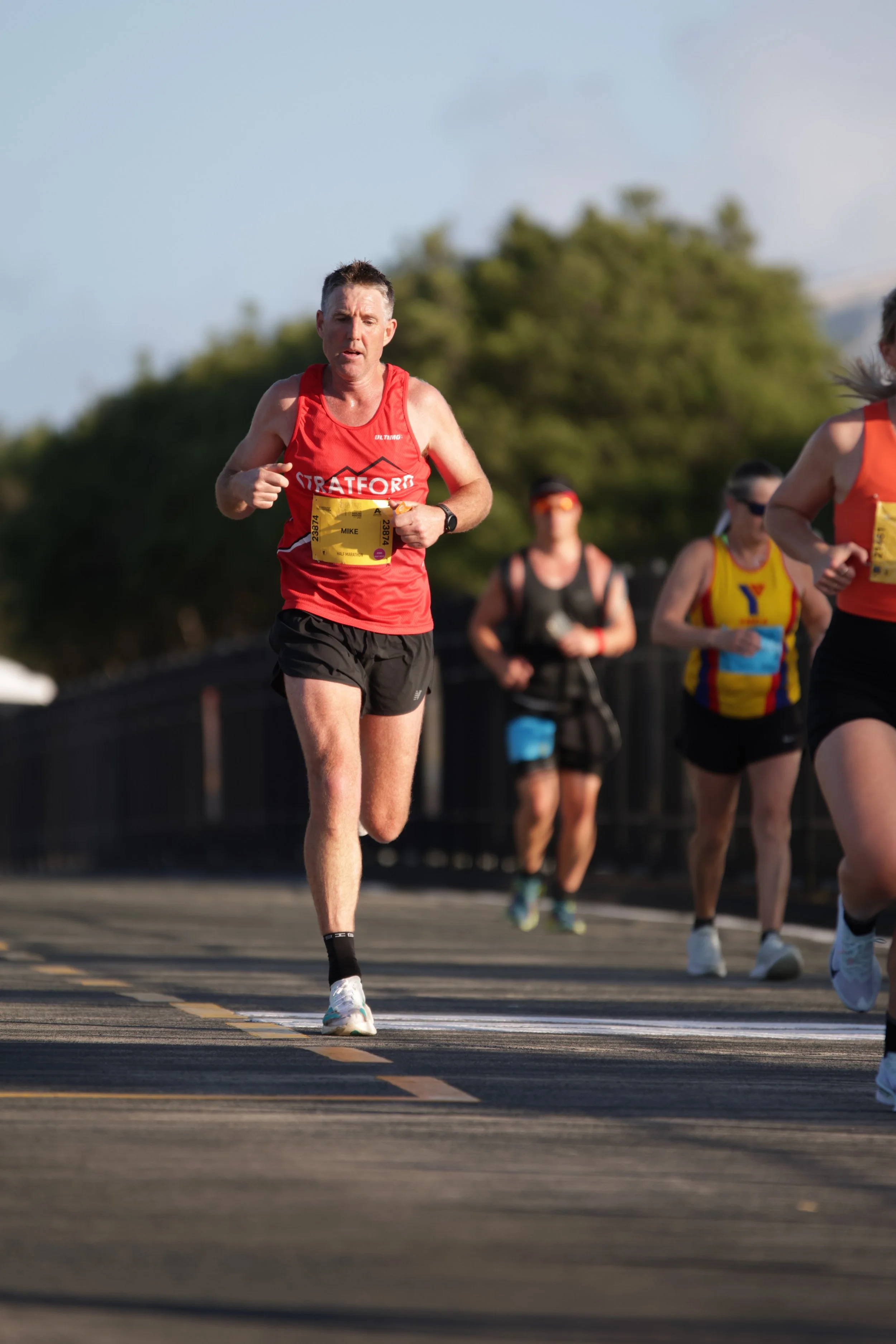 Male runner in red shirt and black shorts participating in a marathon, with other runners in the background on a paved road lined with a black fence and green trees.