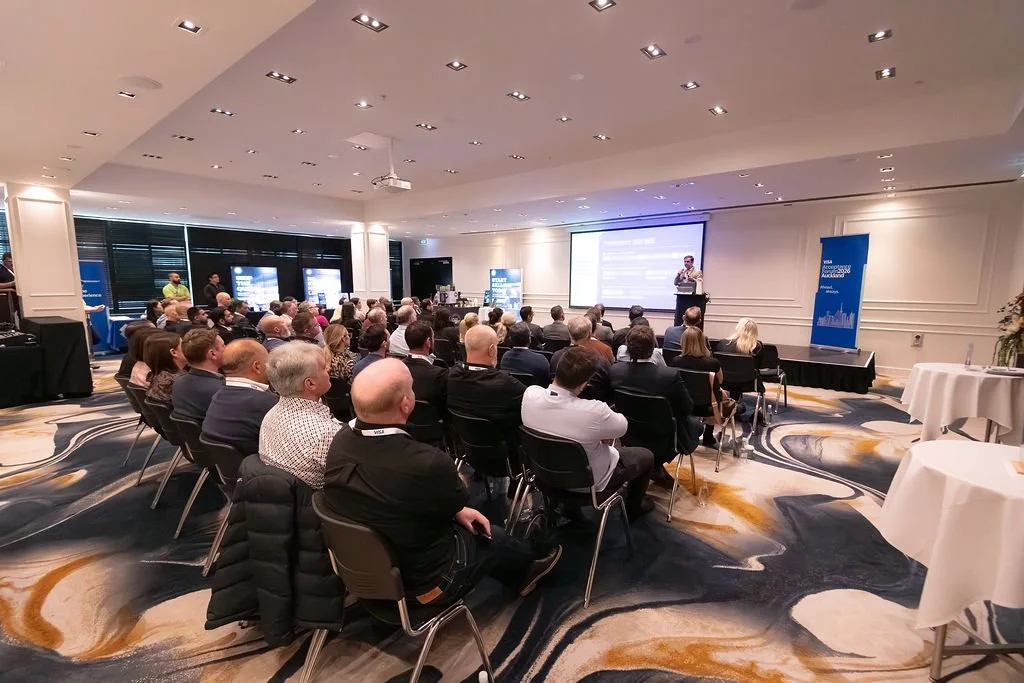 A conference room filled with people seated and listening to a speaker at a podium. Large projection screen displays information behind the speaker, and blue banners are visible on the sides.