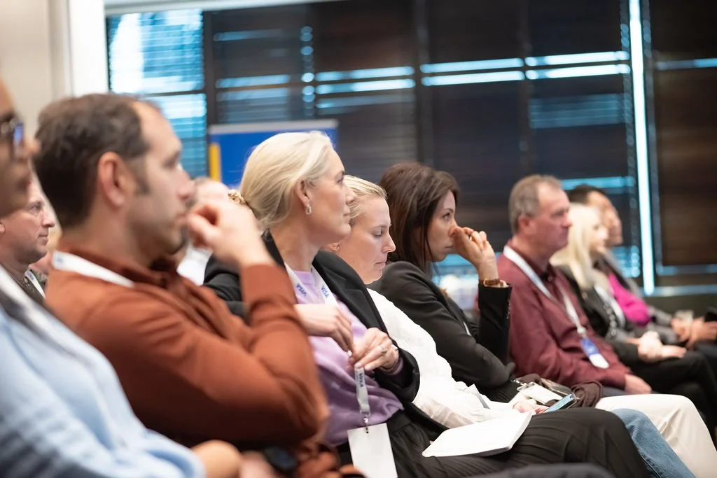Audience seated and attentively listening at a conference or seminar in a room with dark blinds.