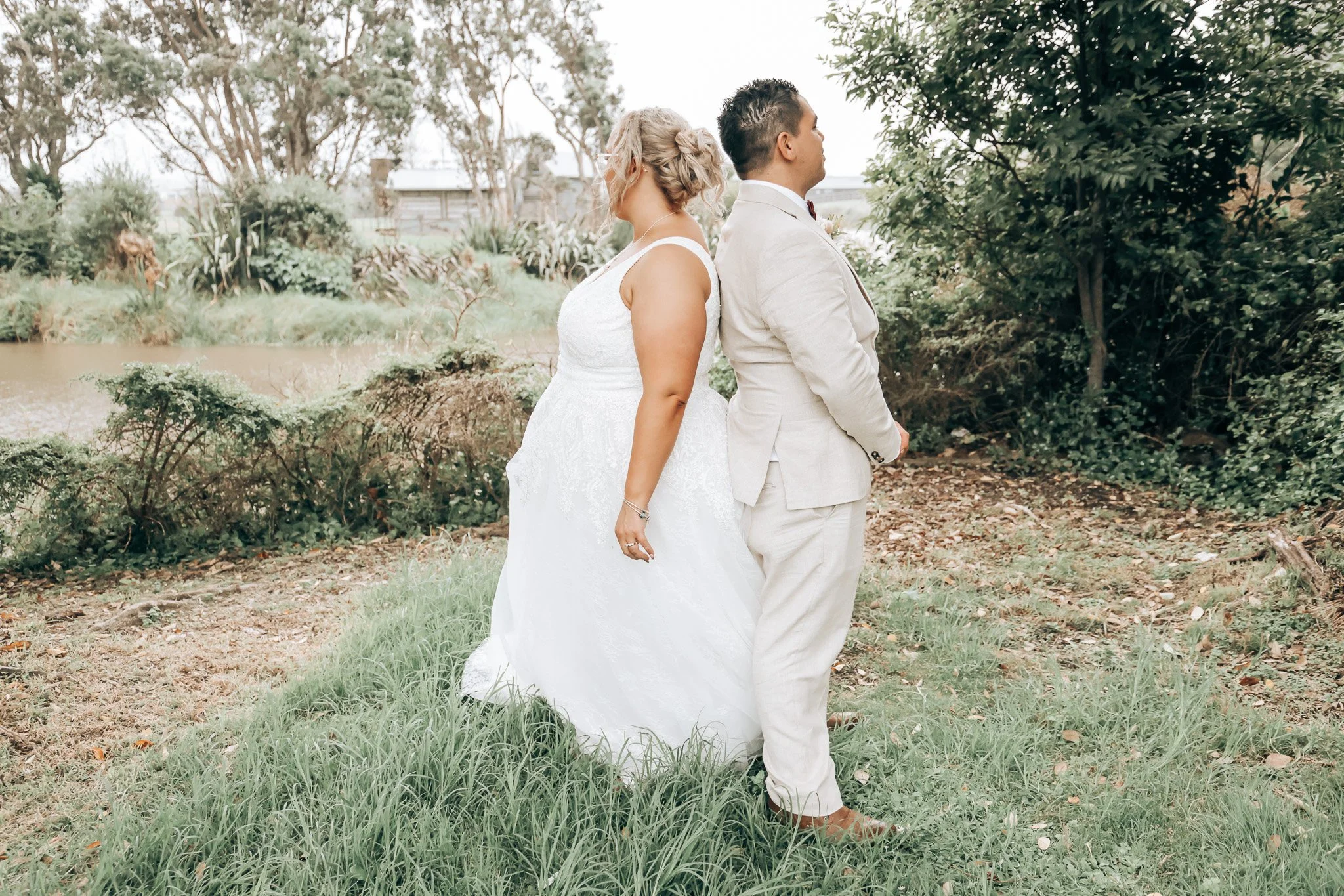 A bride and groom standing back-to-back outdoors among trees and greenery during a wedding ceremony or photoshoot.