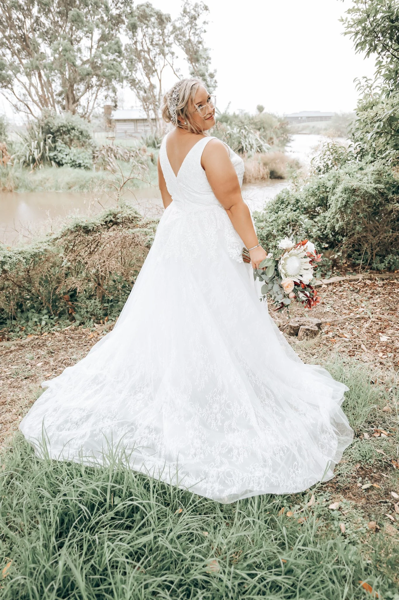 A bride in a white wedding gown holding a bouquet, standing outdoors near a body of water with trees and bushes in the background.