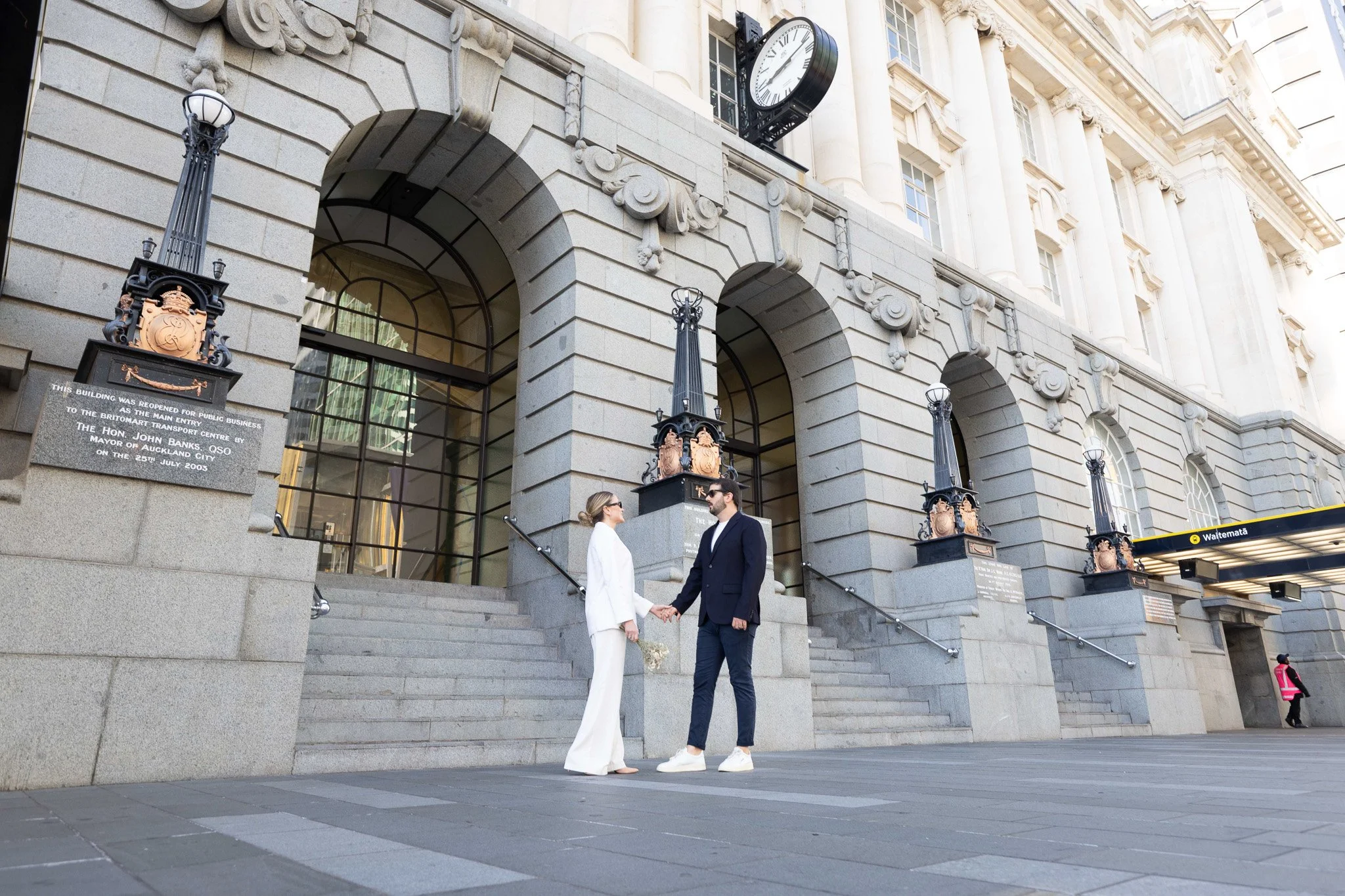 A man and woman in formal attire holding hands and looking at each other on the steps of a historic building with clock and ornate architecture, while a woman in pink stands nearby.