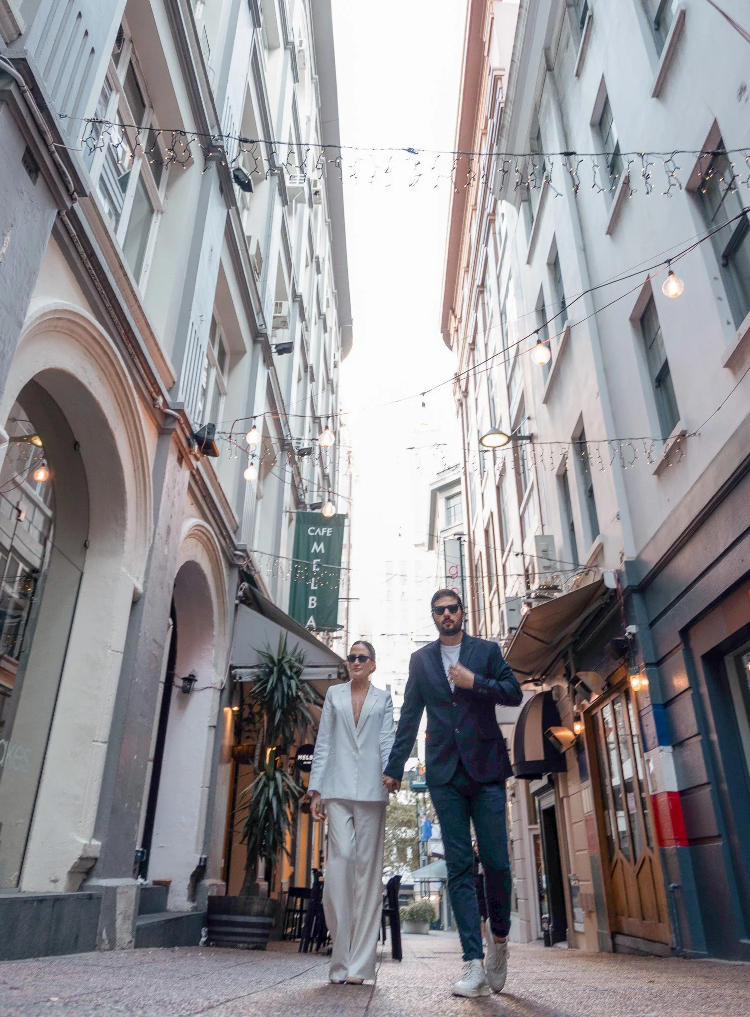 Couple walking hand in hand down a pedestrian street decorated with string lights, surrounded by storefronts and buildings
