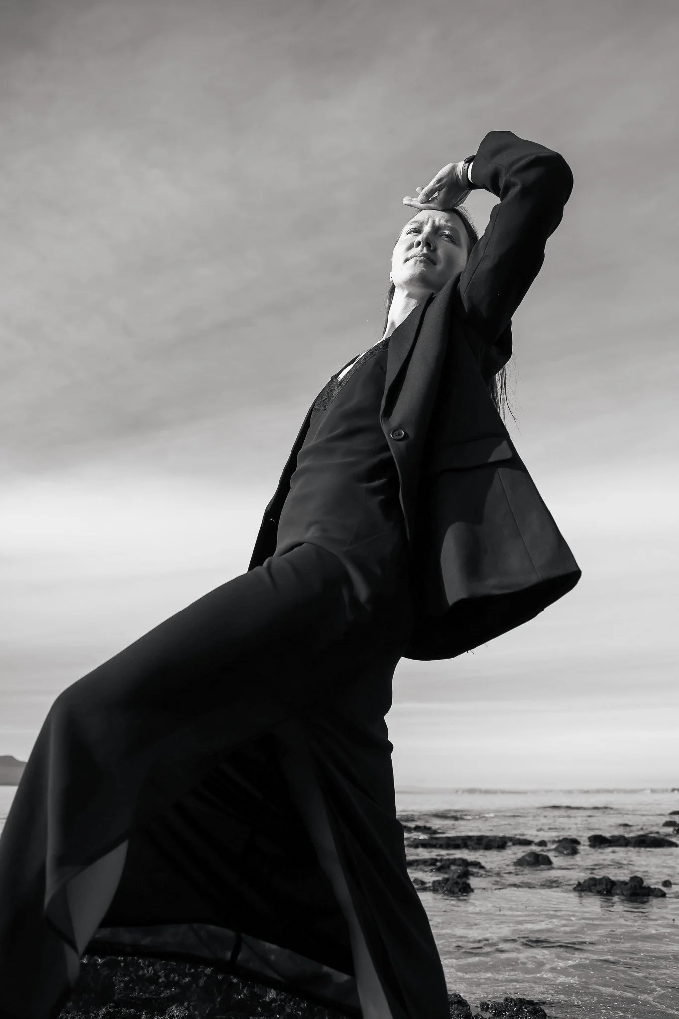 A woman in a black suit standing on a rocky beach, looking up and shielding her eyes from the sun with her hand, sky and ocean in the background.