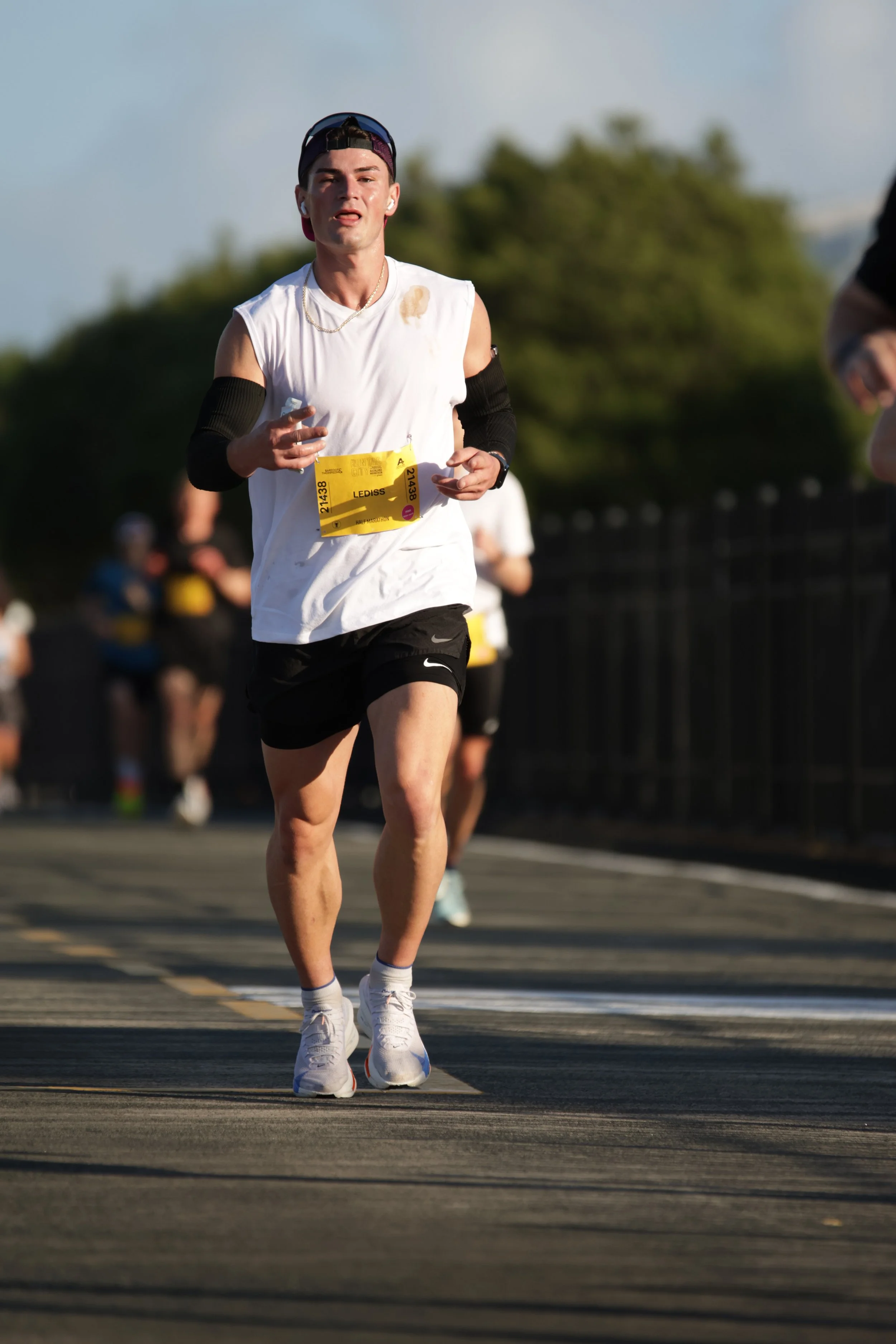 A young man running outdoors during a marathon or race, wearing a white sleeveless shirt, black shorts, white sneakers, and a cap backwards, with other runners in the background.
