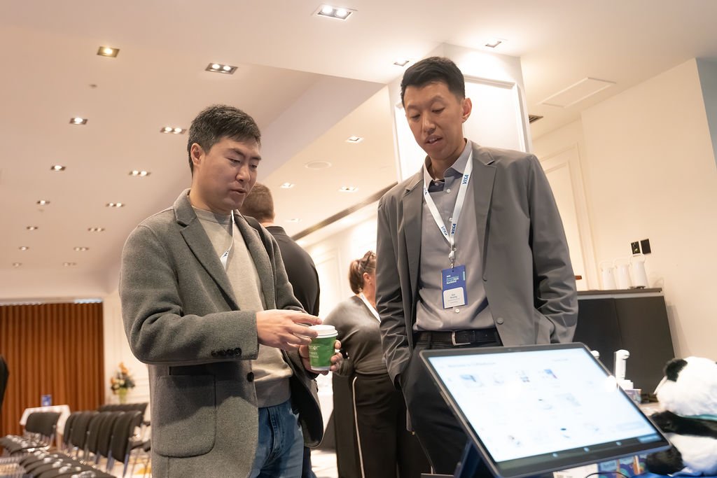 Two men in business attire standing and talking at an indoor conference or business event, with a tablet on a table in front of them.