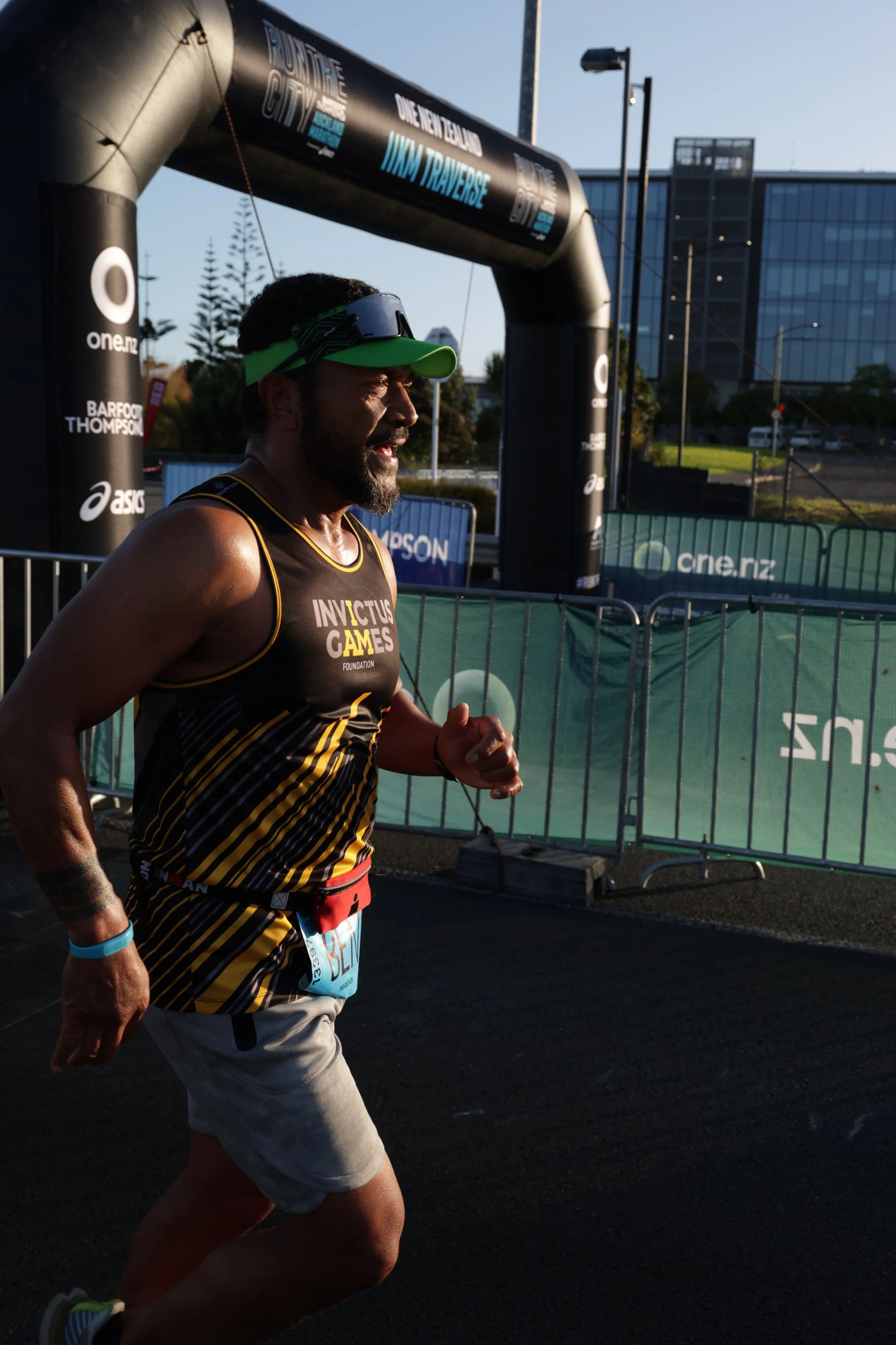 A male runner wearing a green visor, sunglasses, and a black and yellow running outfit, participating in a race outdoors near a start or finish line arch.