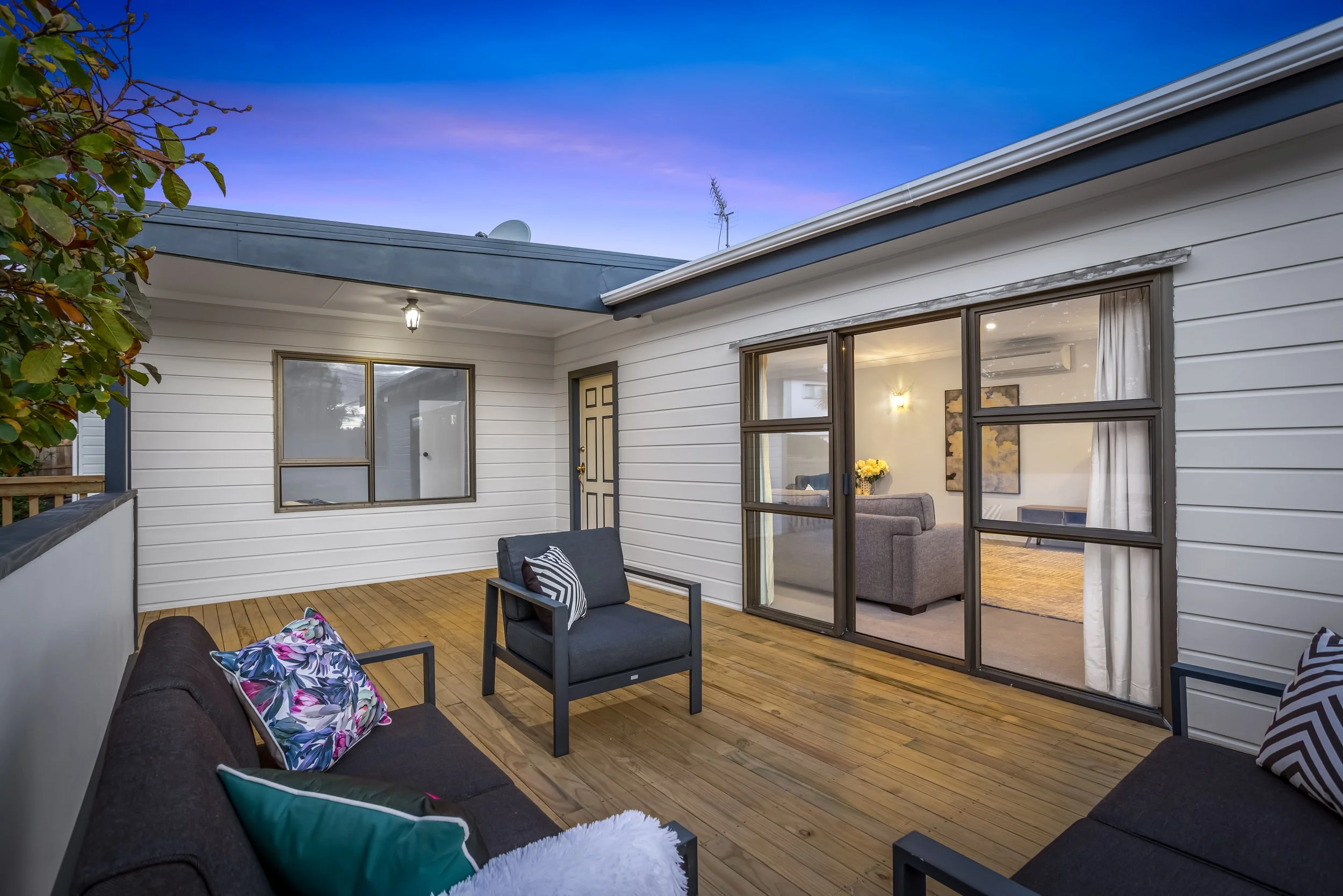 A cozy outdoor balcony space with wooden flooring, black seating furniture with colorful pillows, and view into a warmly lit indoor living room through large sliding glass doors.