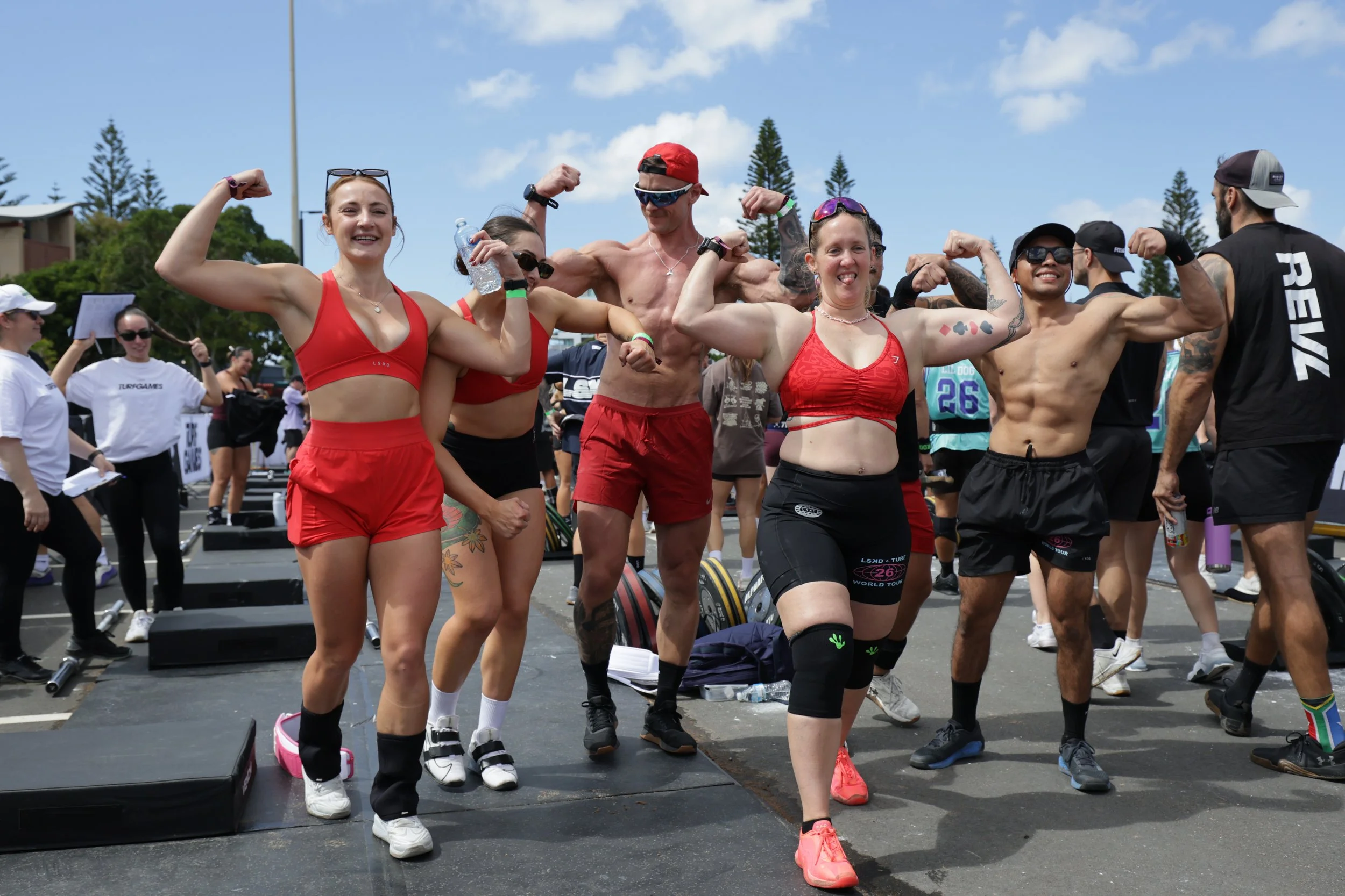 A group of people at an outdoor fitness event, with some flexing their muscles and smiling, dressed in athletic attire, including tanks, shorts, and sunglasses, with gym equipment and a crowd in the background.