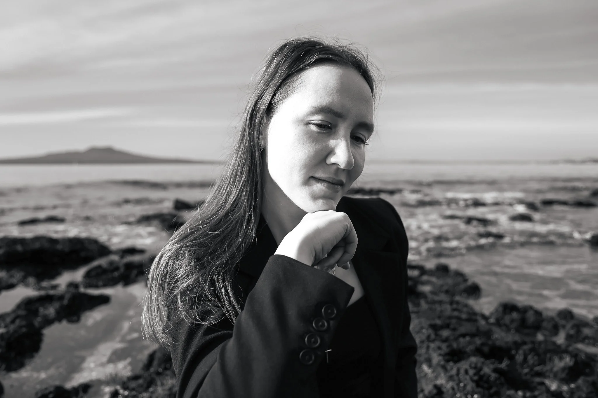 A woman with long hair, wearing a dark jacket, is standing outdoors near a rocky shoreline with water and distant land in the background, captured in black and white.