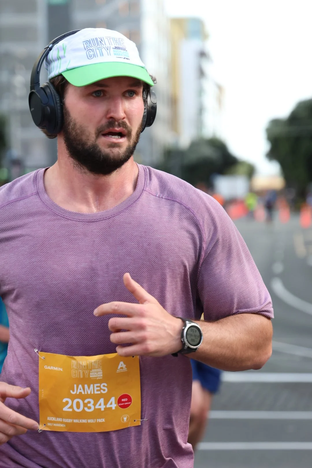 A man running in a race wearing a purple shirt, a cap that says 'Run the City,' a wearable watch, and a yellow race bib with the name 'James' and bib number 20344.