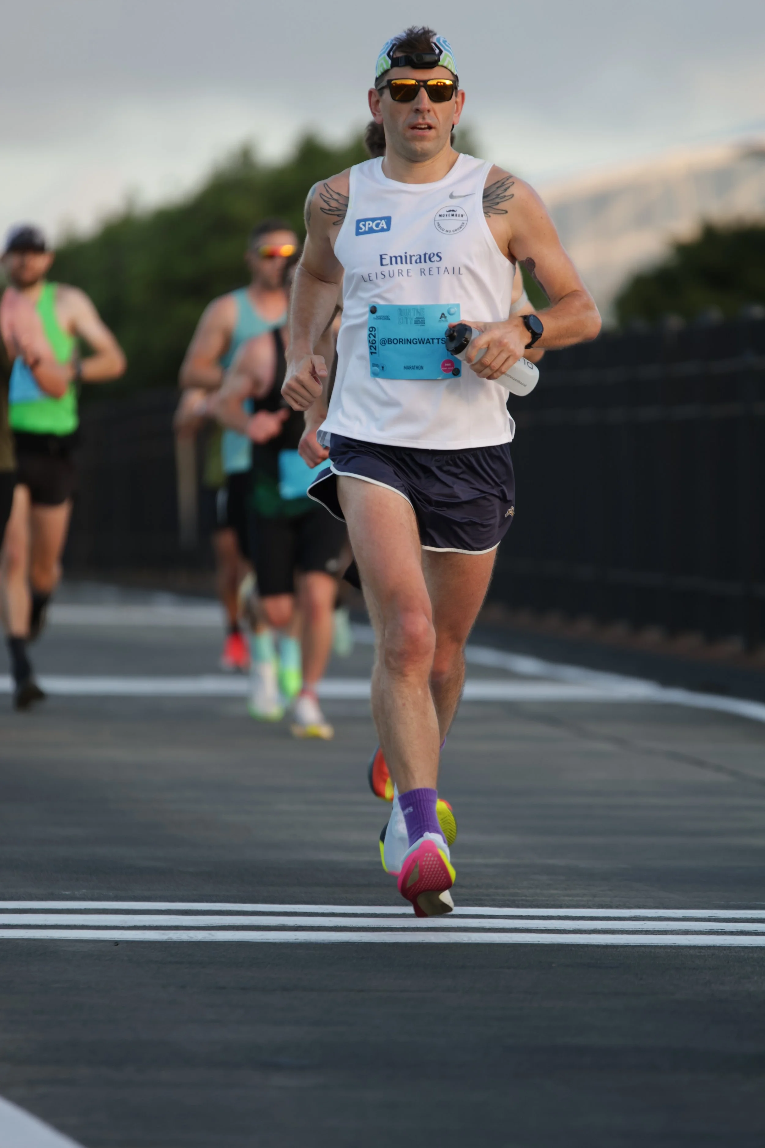 A male marathon runner in a white tank top, black shorts, and colorful running shoes, holding a water bottle, running on a road during a race with other runners in the background.