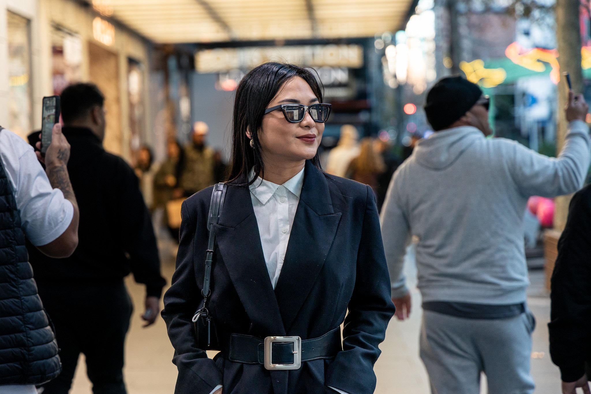 Woman with dark hair, wearing sunglasses, a navy blazer, and a white shirt, standing among a crowd outdoors.