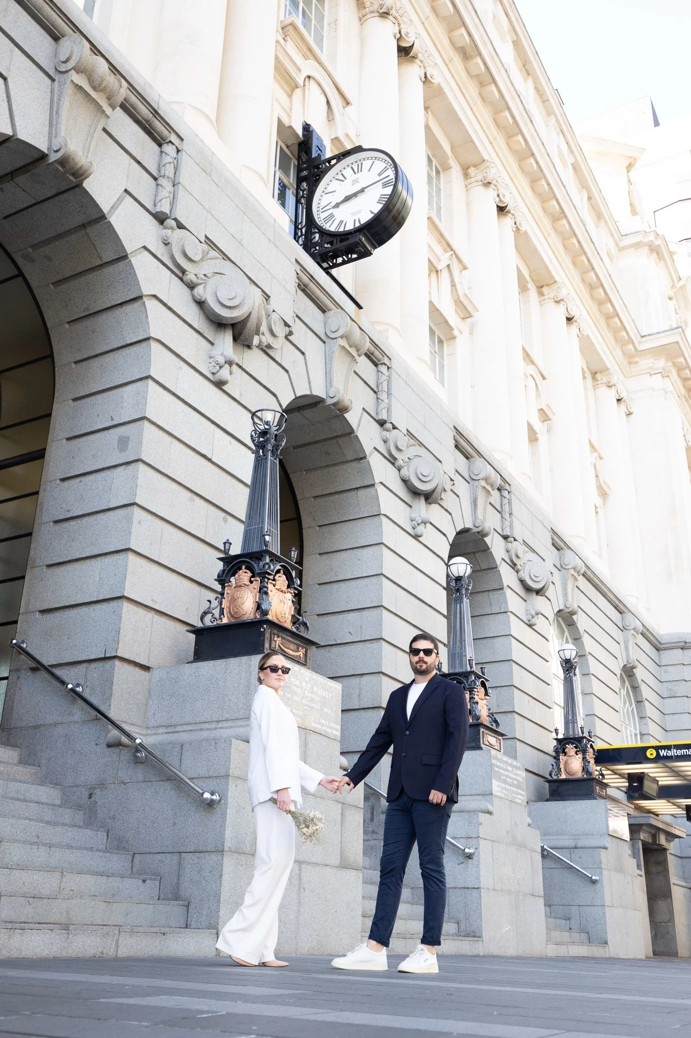 A couple dressed in formal attire holding hands on the steps of a grand historic building with a clock, decorative columns, and street lamps.