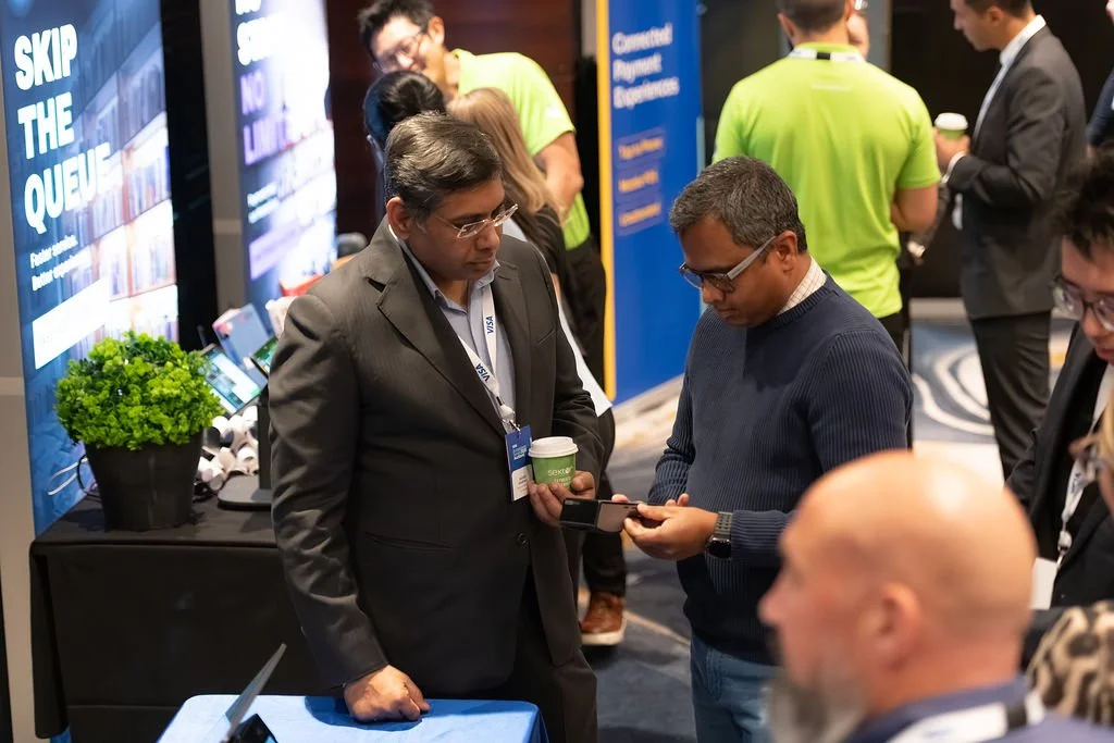 Two men, one in a suit and the other in a sweater, are looking at a smartphone at a conference. The man in the suit is holding a coffee cup, and there are other people and display booths in the background.