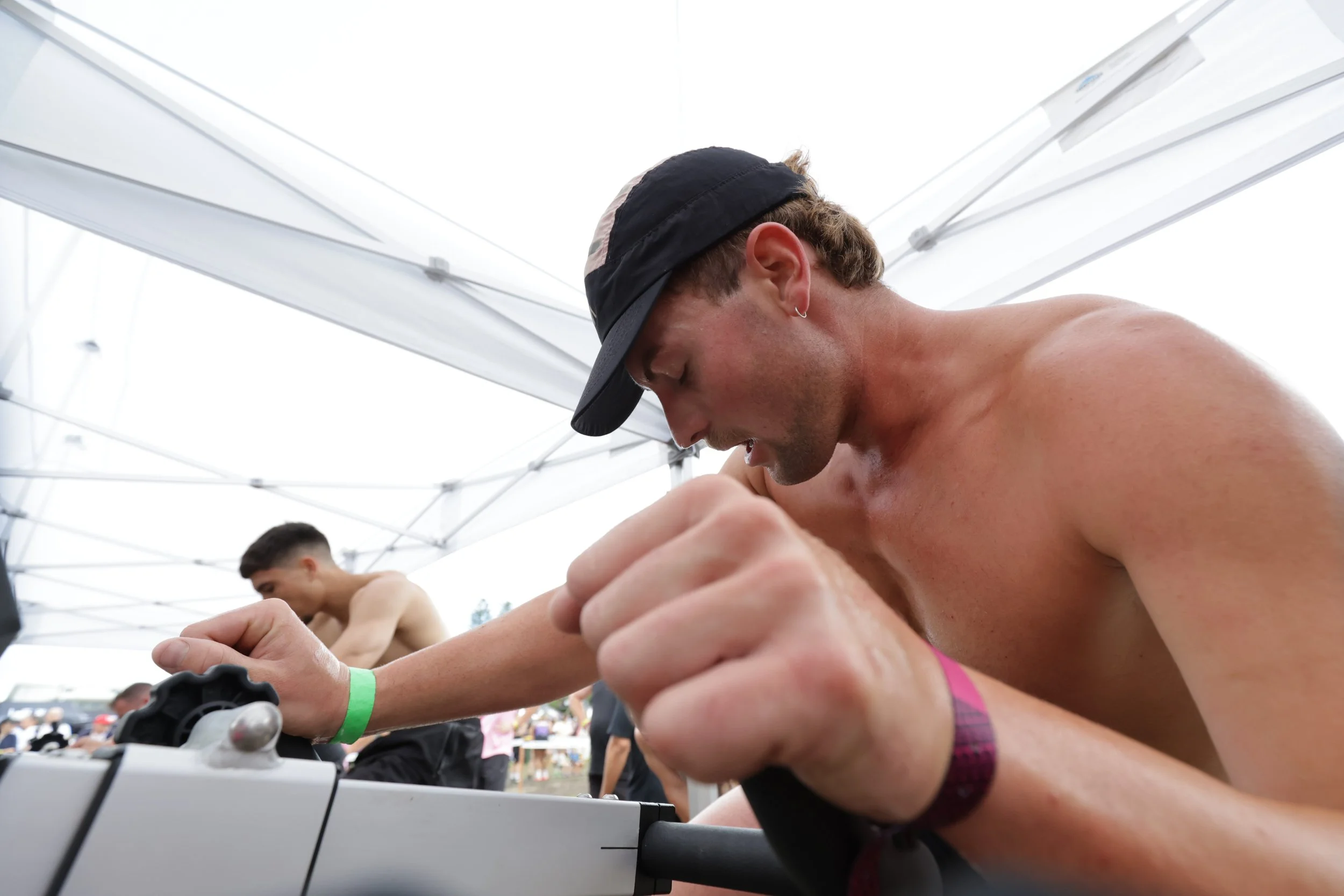 A man wearing a black cap and a pink plaid shirt with rolled sleeves is playing an electronic keyboard at an outdoor event under a white canopy. There are other people in the background, including another shirtless man, and the event appears lively.