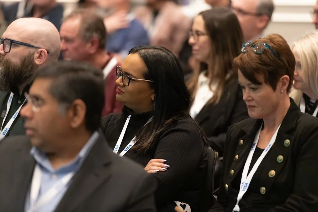 Attendees sitting in a conference room, some with lanyards, listening attentively.