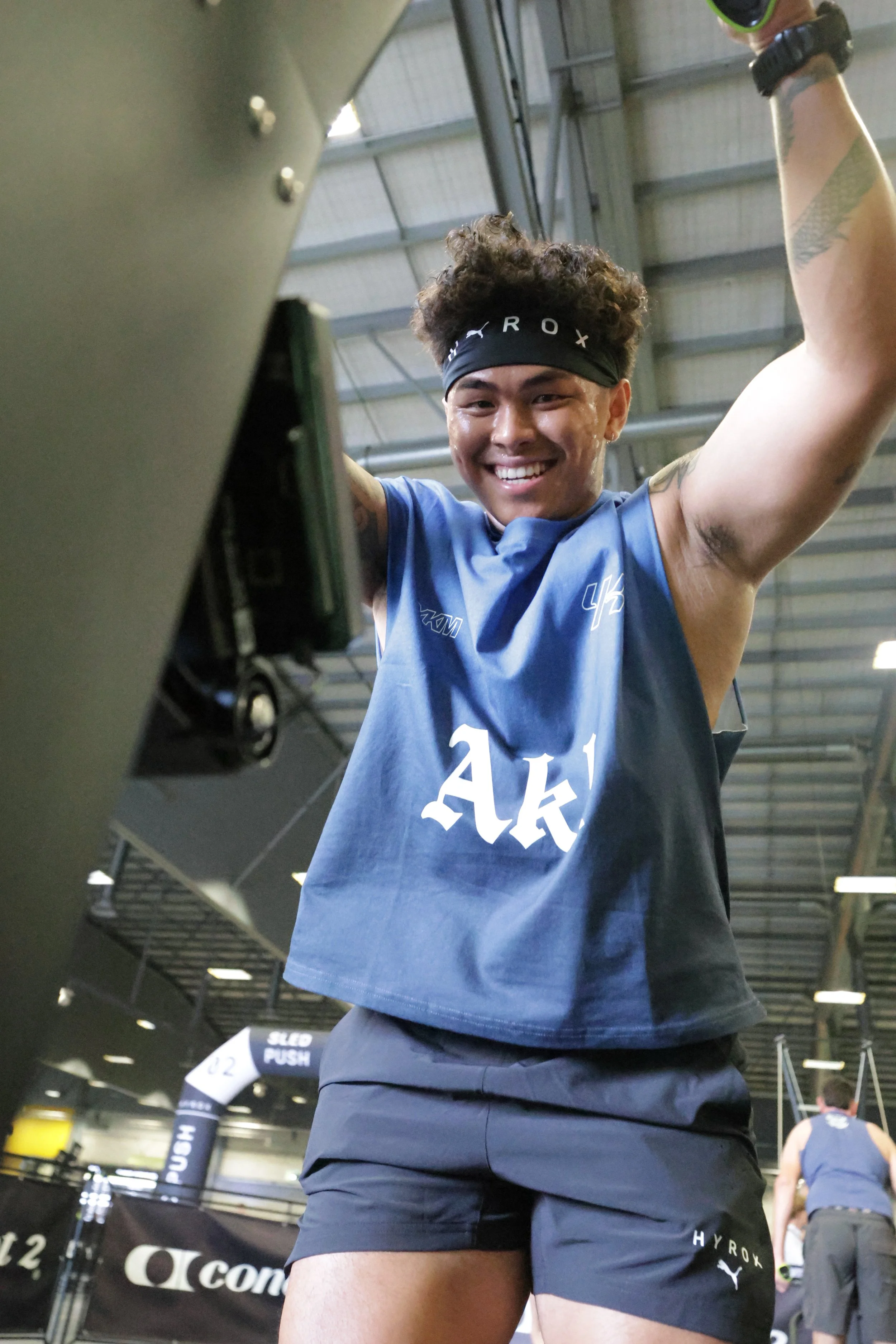 A young man with curly hair, wearing a black headband, blue sports jersey, and black shorts, smiling and raising his arms in a gym.