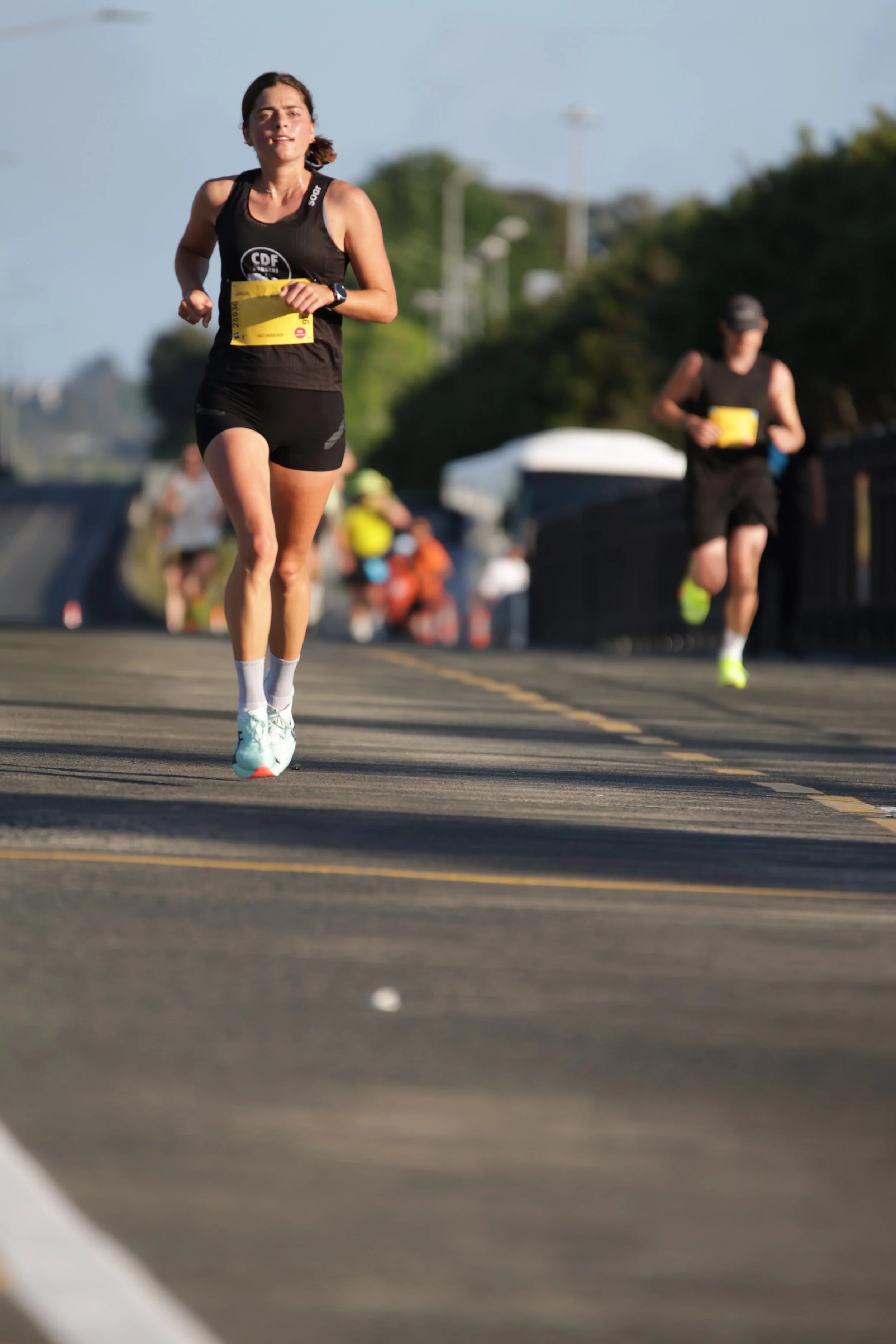 A woman running in a race, wearing a black athletic outfit with a yellow bib, on a road with a blurred background of trees and other runners.