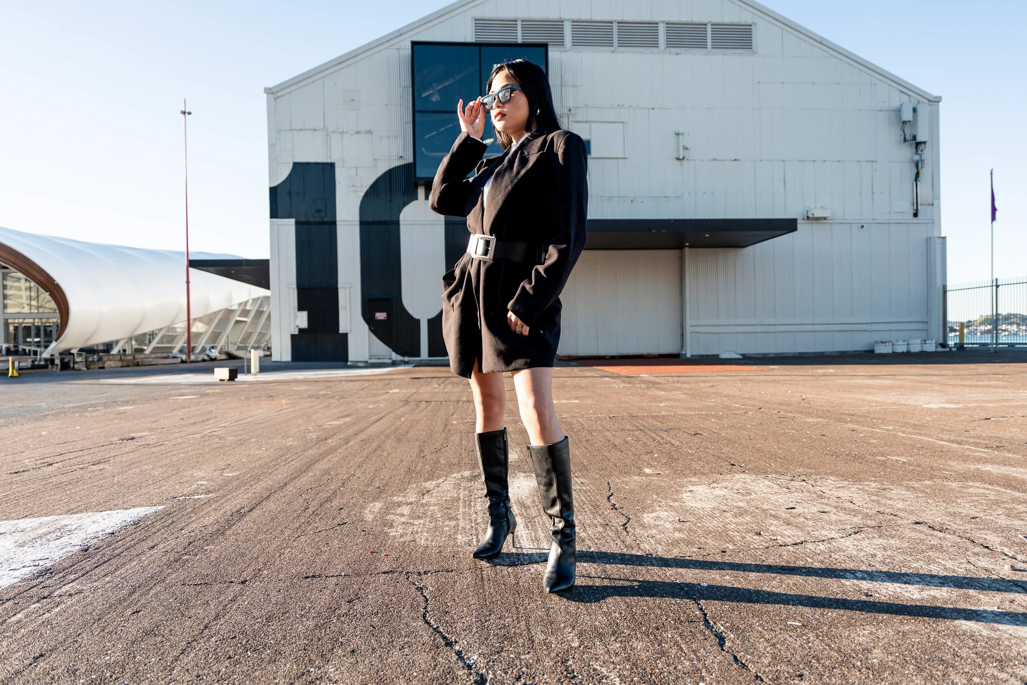 A woman in a black coat and high-heeled black boots standing outdoors in a parking lot, adjusting black sunglasses, with a large white building and clear blue sky in the background.