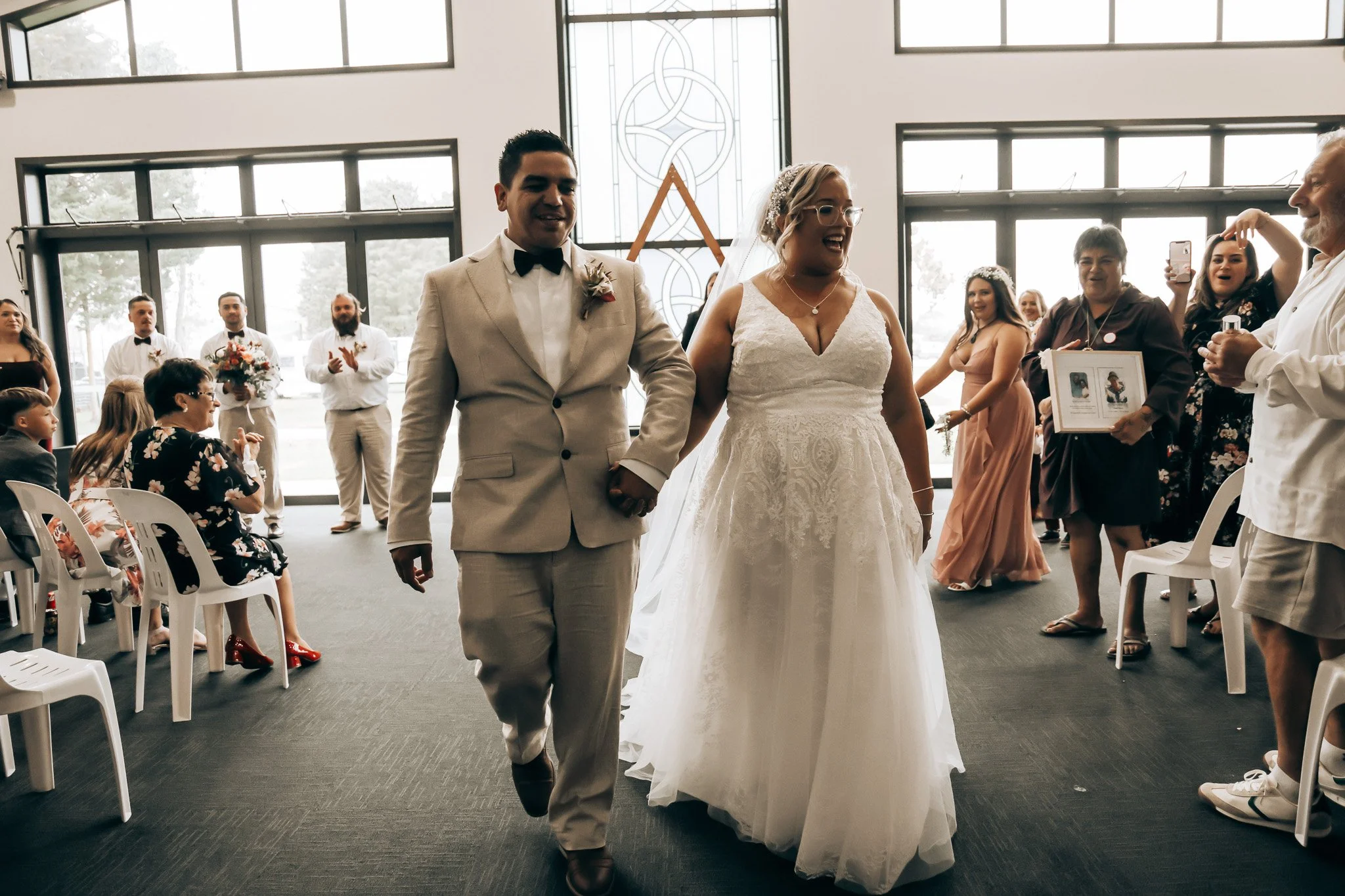 A bride and groom walking hand in hand during their wedding ceremony inside a modern chapel with large windows and stained glass door, surrounded by guests.